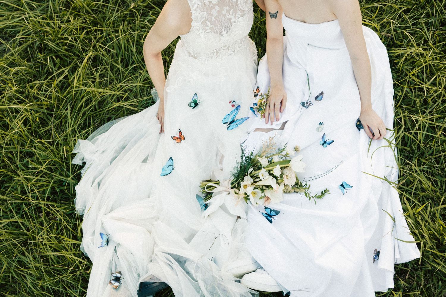 Overhead view of two brides lying in grass with blue butterflies on their white gowns.