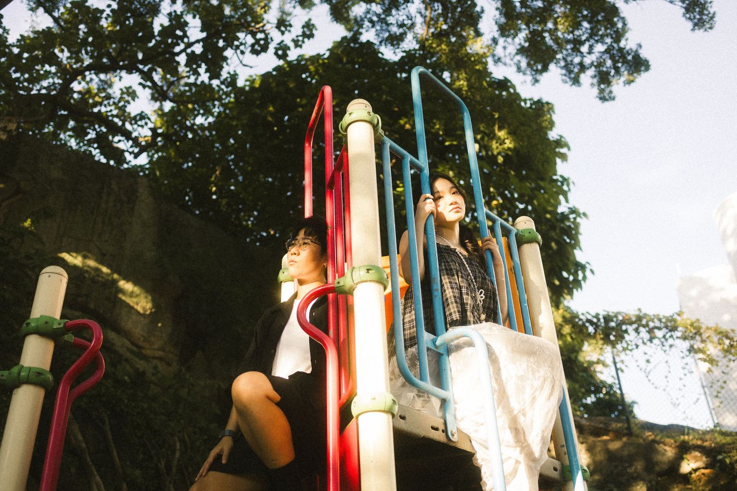 Two young people standing on colorful playground climbing structure under shady trees.