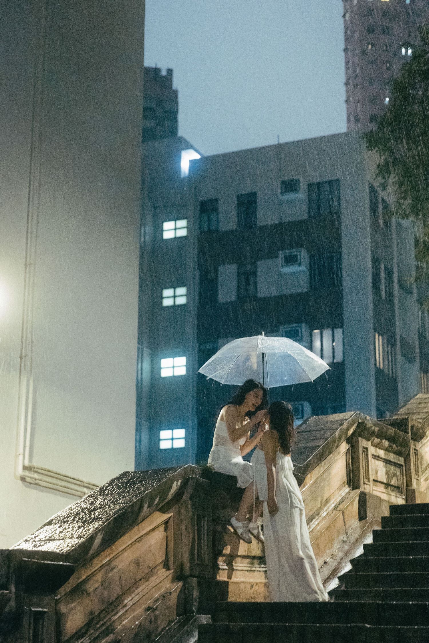 Two women in white dresses sharing umbrella on rainy night city steps.