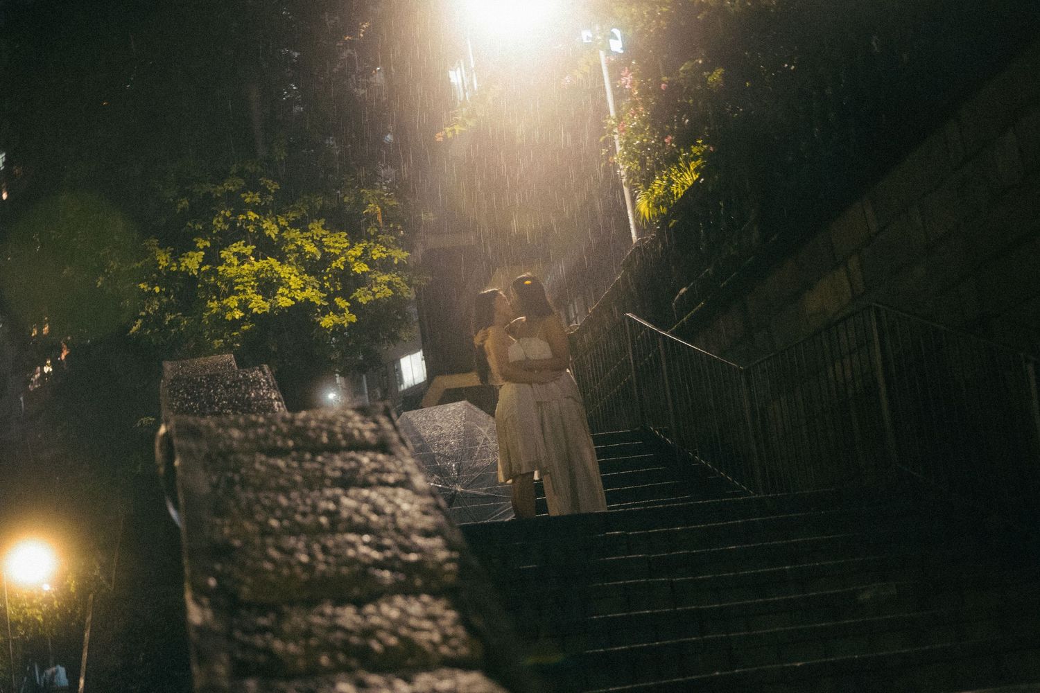 Two women in white dresses embrace on stone steps under dramatic night lighting.