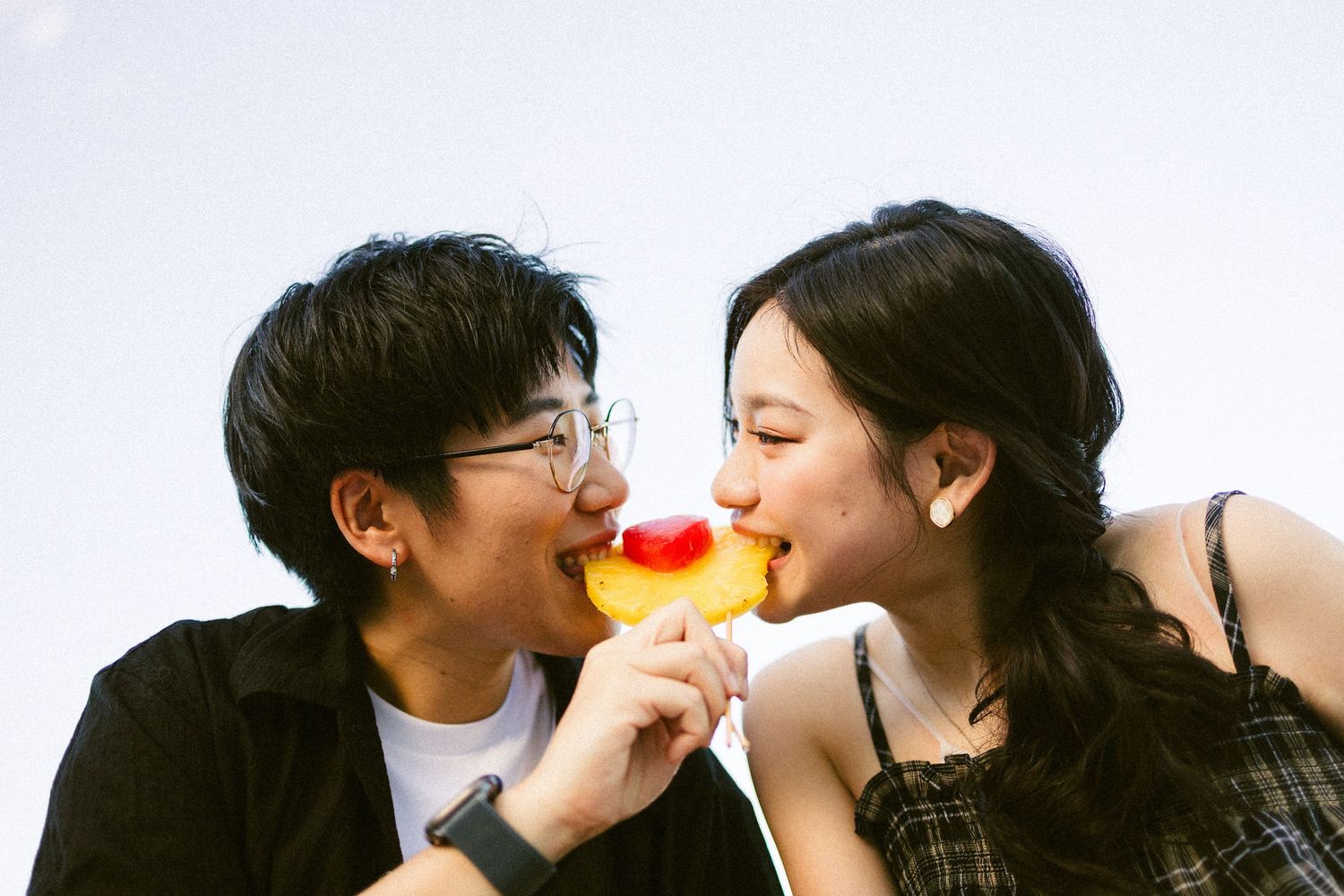 Couple sharing a fruit popsicle together against a bright sky, smiling joyfully.
