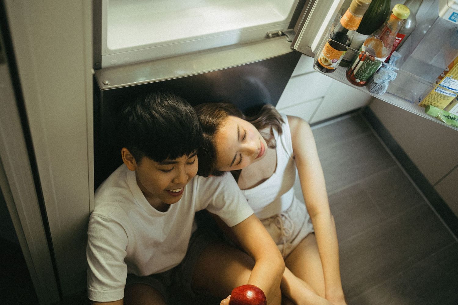 Young couple sitting on kitchen floor laughing together in front of open refrigerator.