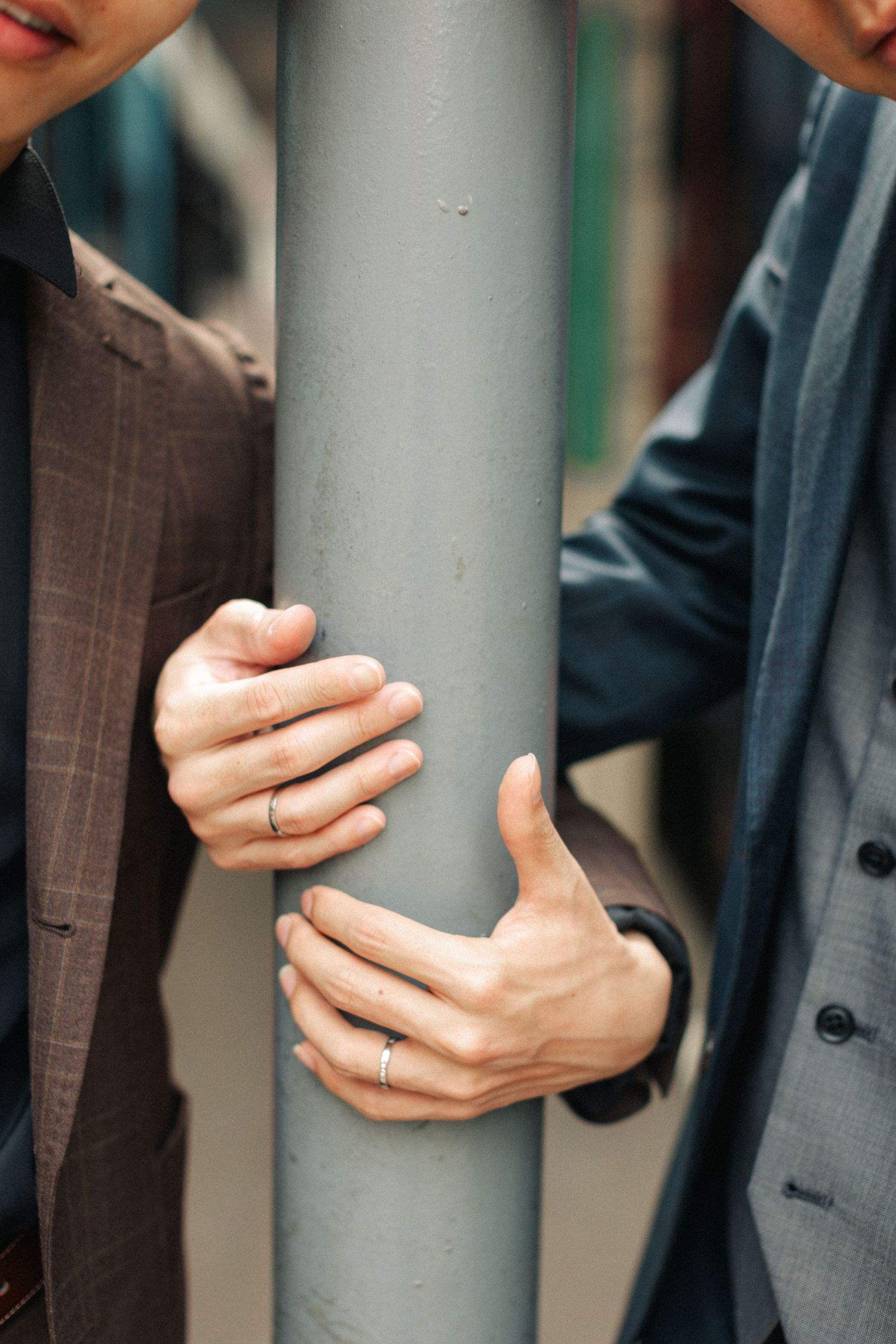 Close-up of two men in suits with hands touching a grey pole, both wearing rings.