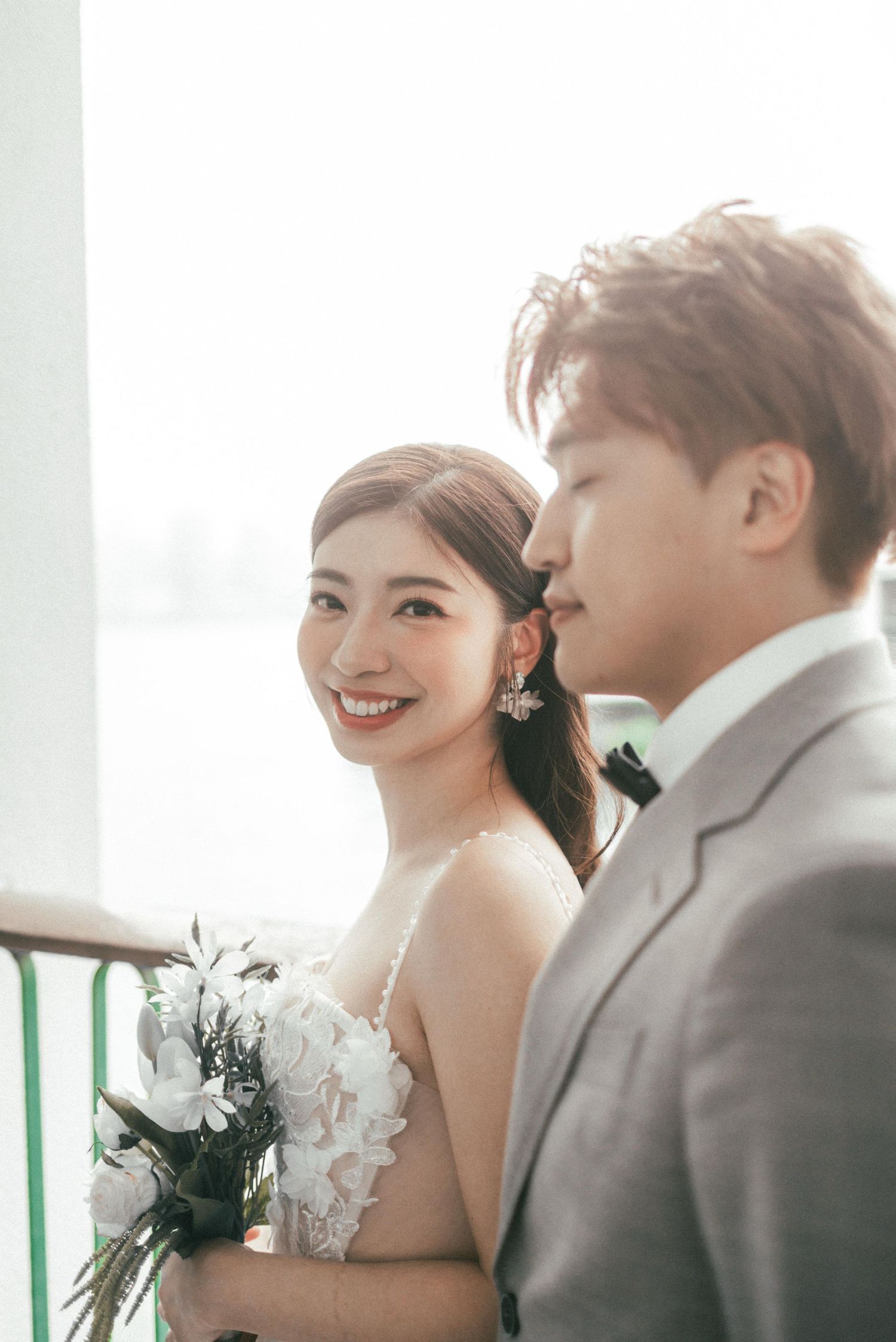 Smiling Asian bride in a white gown holding white flowers beside a groom in a grey suit.