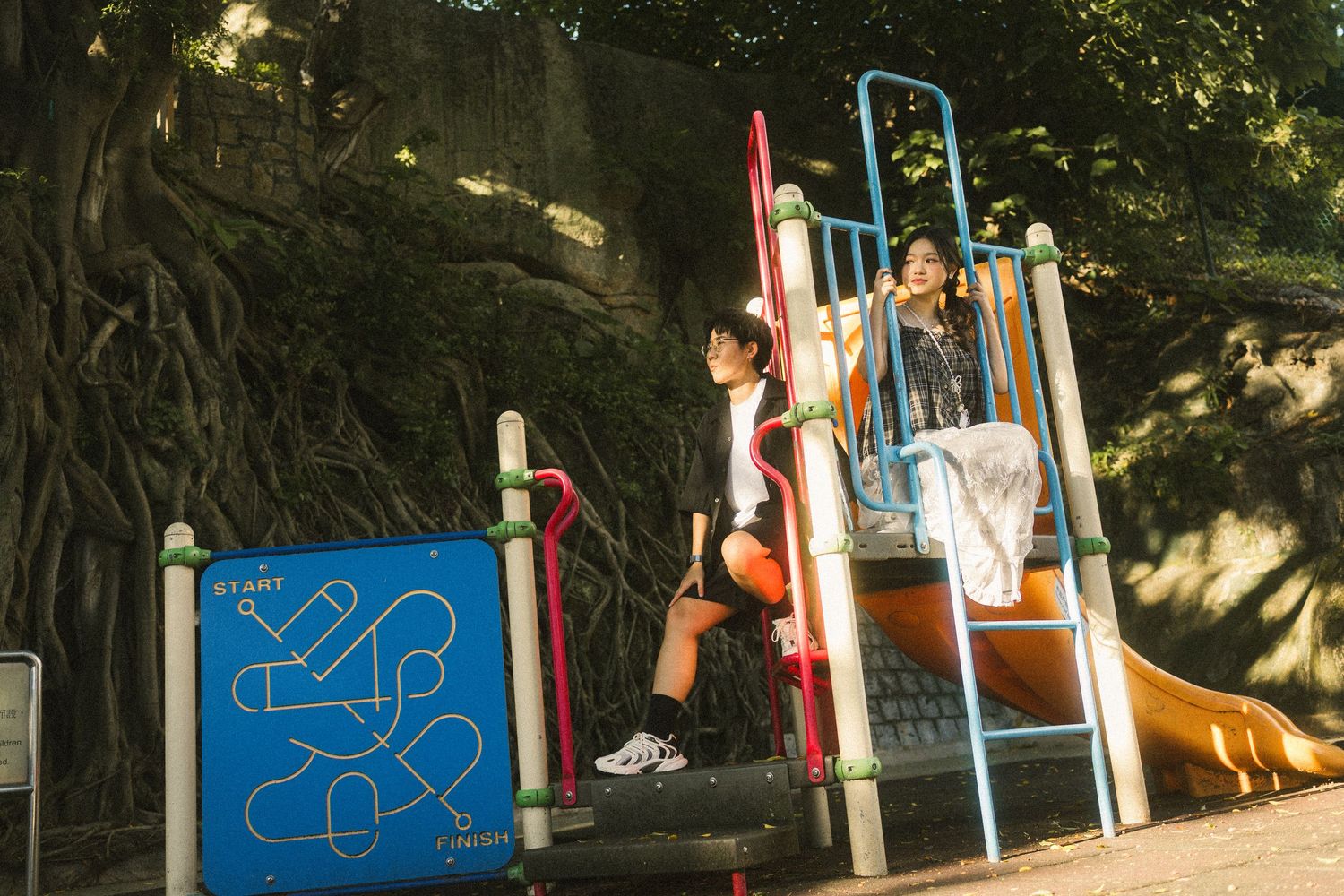 Two children playing on a colorful playground structure with a large tree in the background.