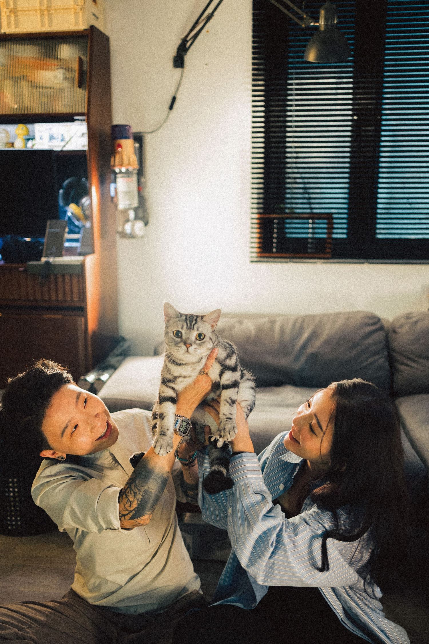 Couple playfully lifting a silver tabby cat together in a cozy apartment living room at night.
