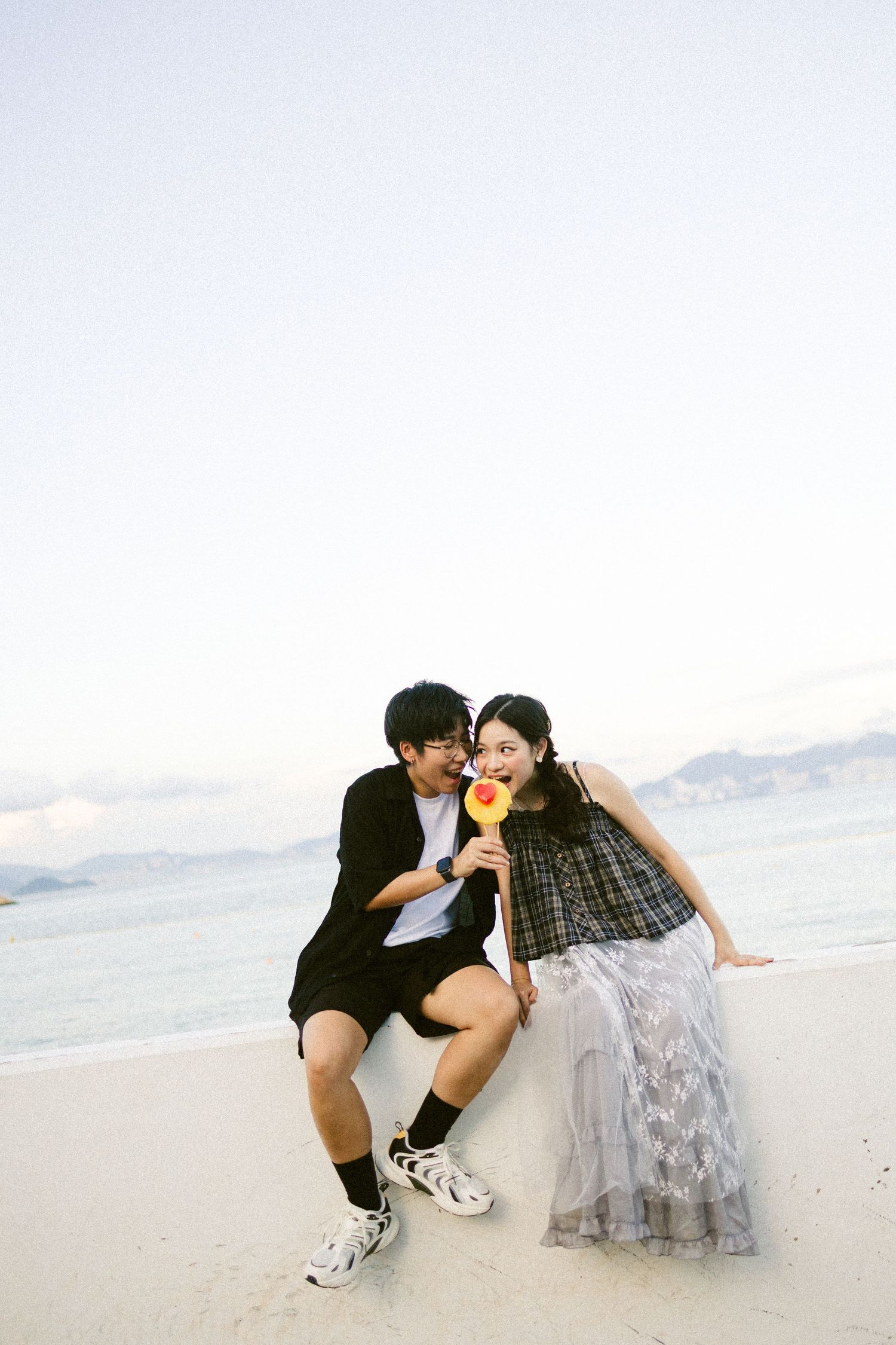 Couple sitting on a sandy beach at dusk sharing a colorful lollipop by the ocean.