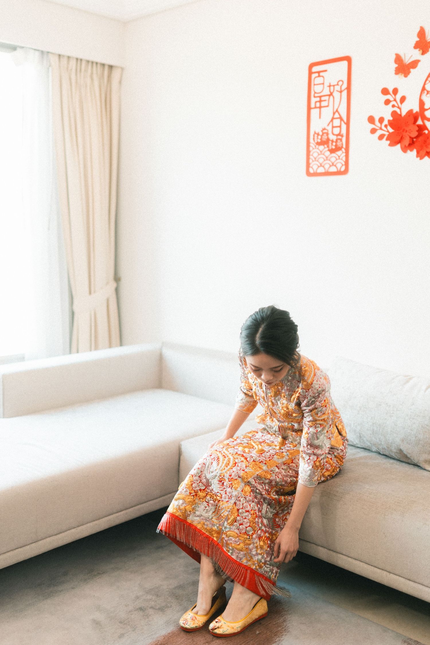 Bride in vibrant orange floral qipao sits on white sofa adjusting golden embroidered shoes during wedding preparations.