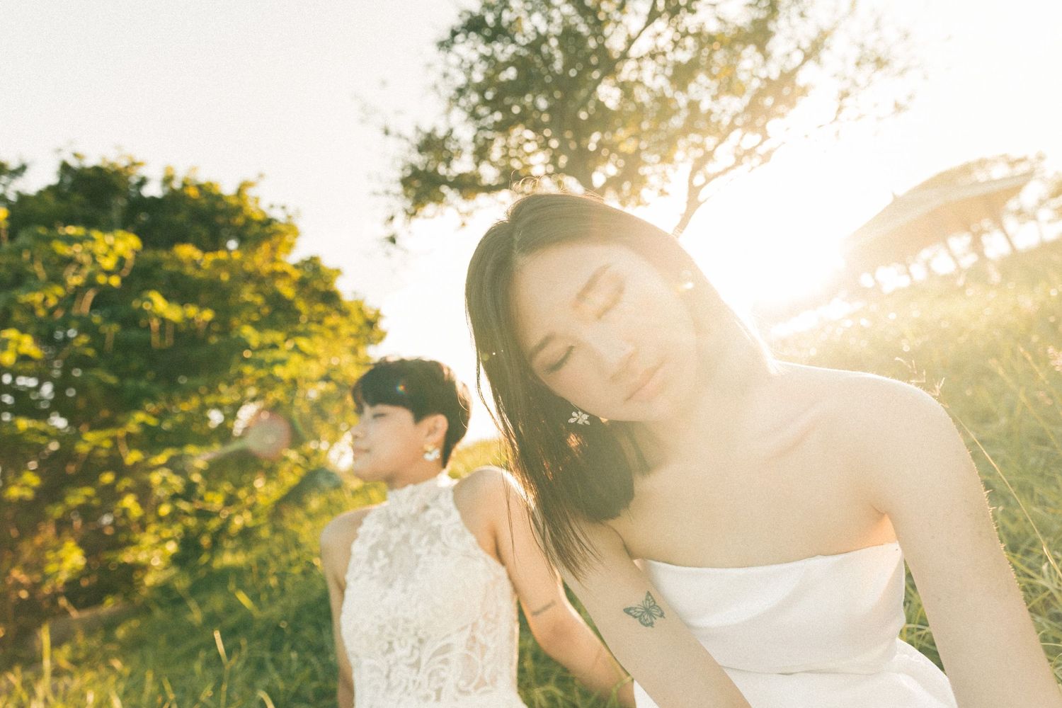 Two brides in white gowns share a romantic moment in a golden sunlit outdoor setting.