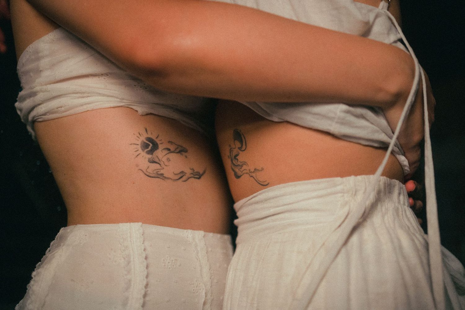 Two women in white dresses embrace, showing matching floral tattoos on their sides.