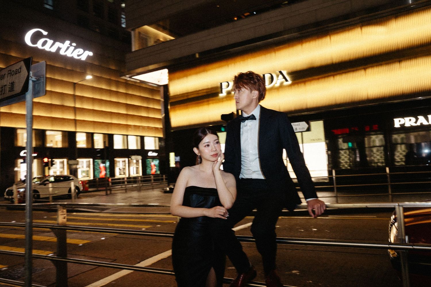 An elegant couple in formal wear poses on a city street near luxury boutiques at night.