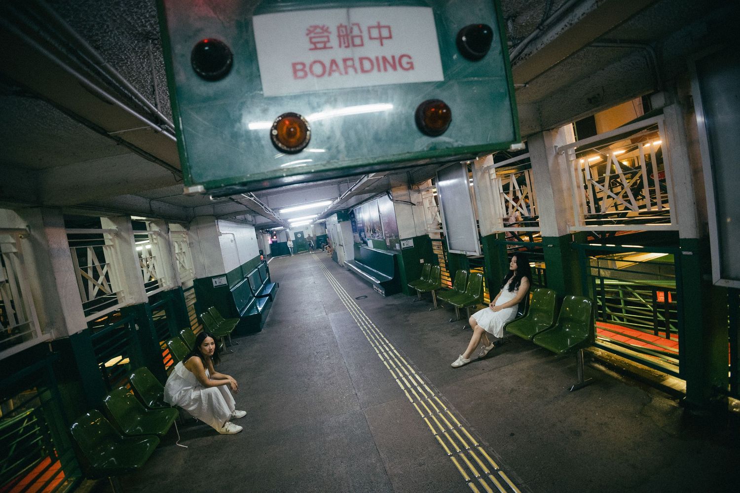 Two women in white dresses sit alone in a dimly lit ferry boarding area.