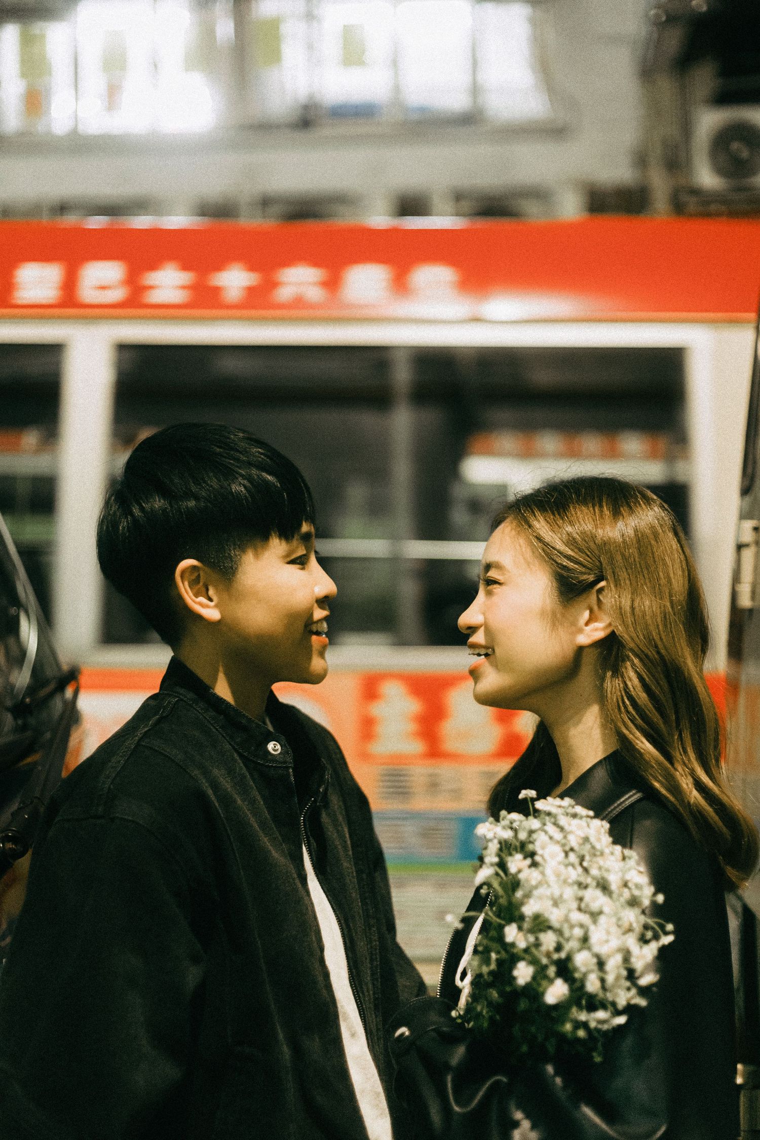 A smiling Asian couple facing each other at night, with the woman holding white flowers.