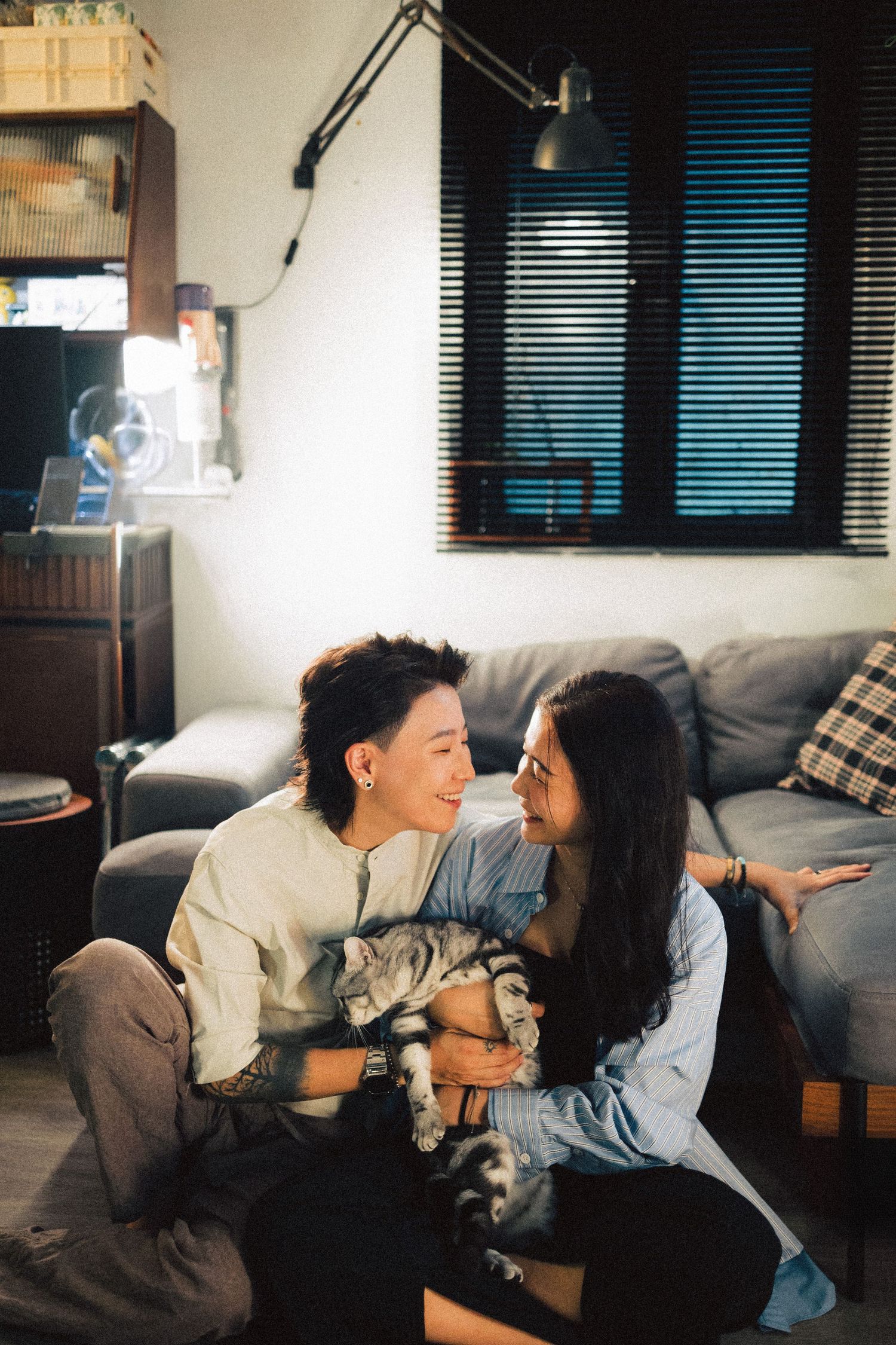 Two young Asian women smiling at each other while holding a tabby cat in a cozy living room.