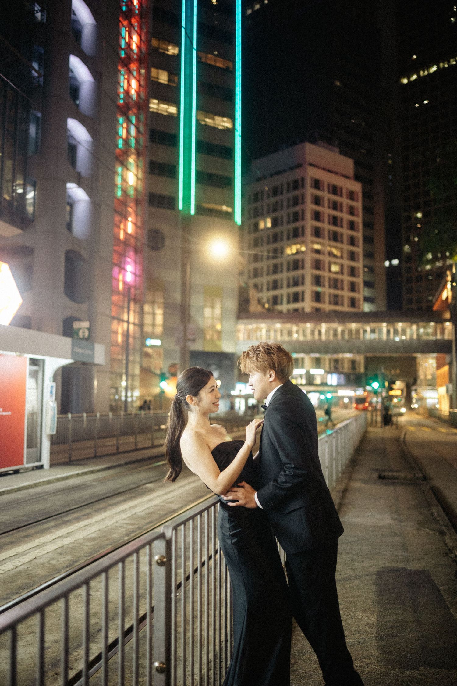 A couple in formal black attire embraces romantically on a city bridge at night.