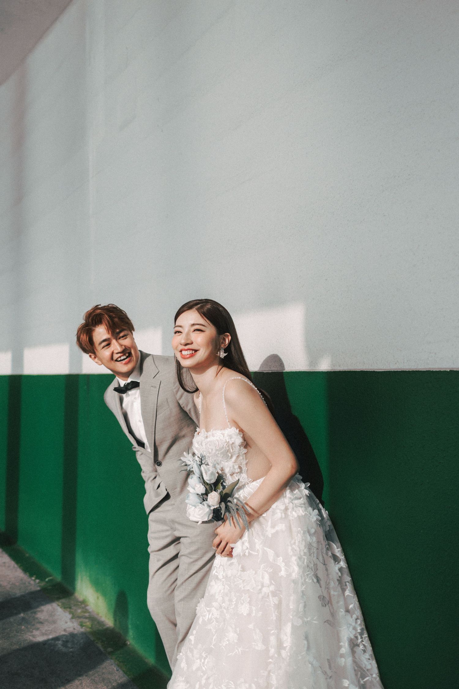 Smiling bride and groom pose against a green and white wall in wedding attire with bouquet.