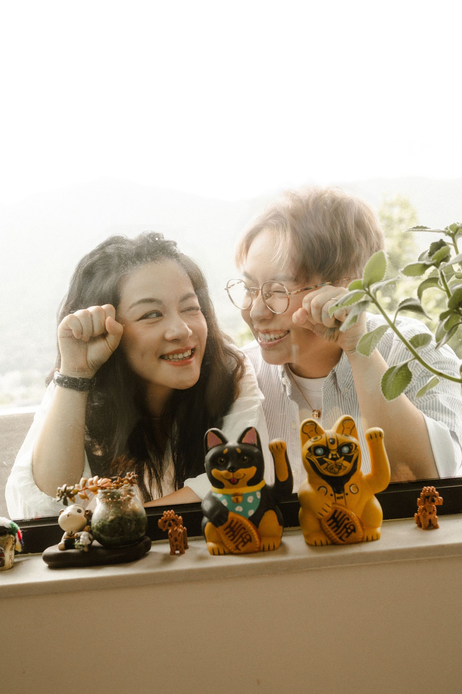 Happy couple smiling together beside lucky cat figurines on a sunlit windowsill with green plants nearby.