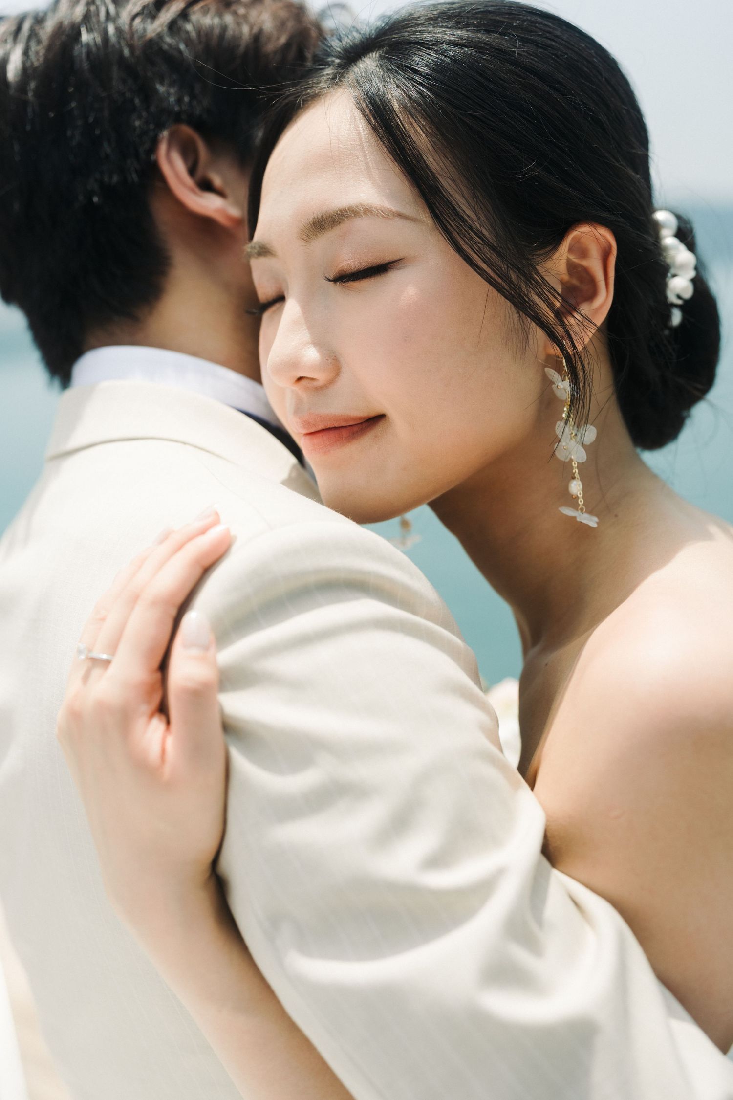Asian bride with pearl hair accessories smiles contentedly while embracing groom in cream suit outdoors.