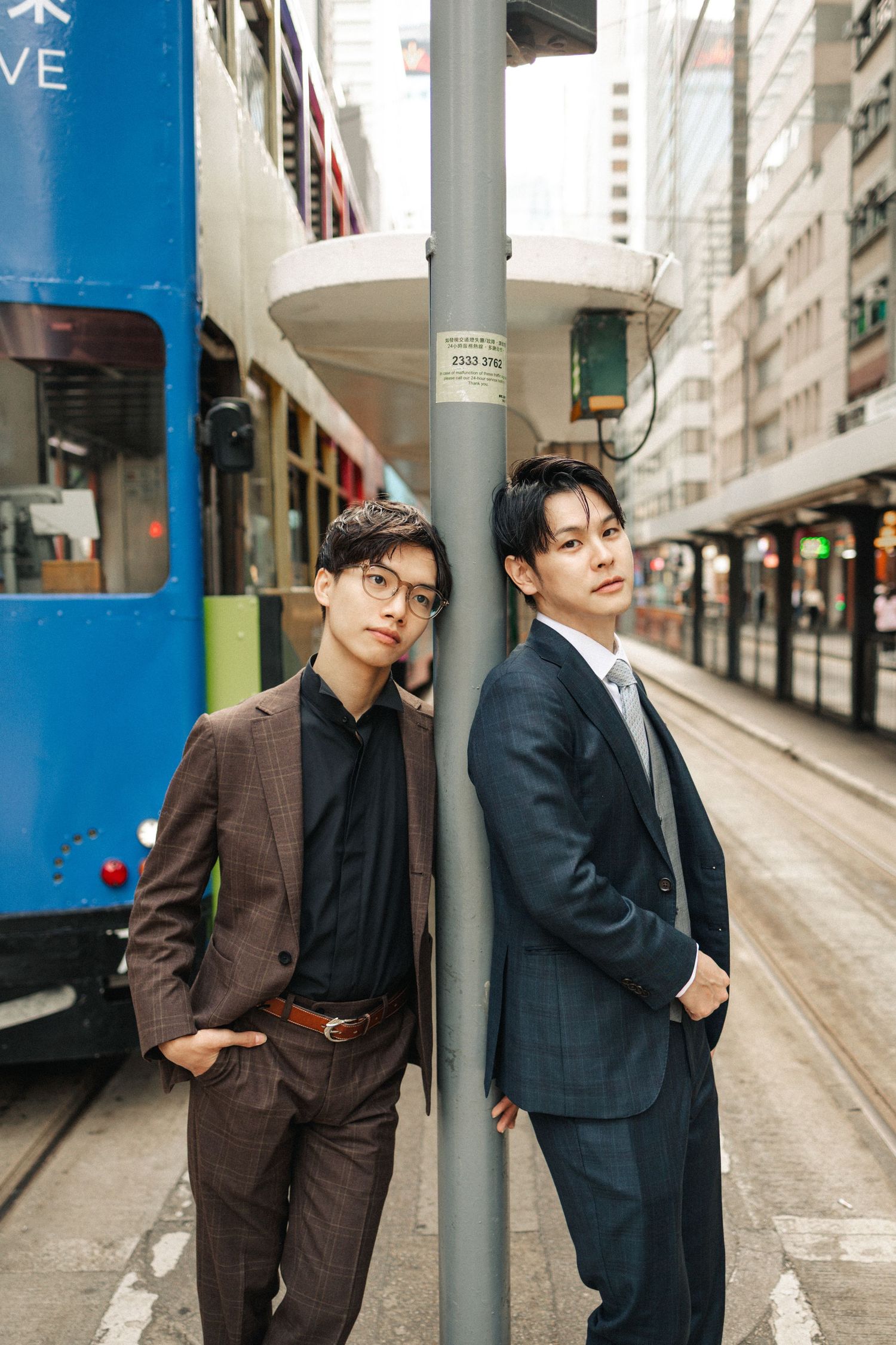 Two stylish Asian men in suits lean against a tram stop pole on a Hong Kong street.