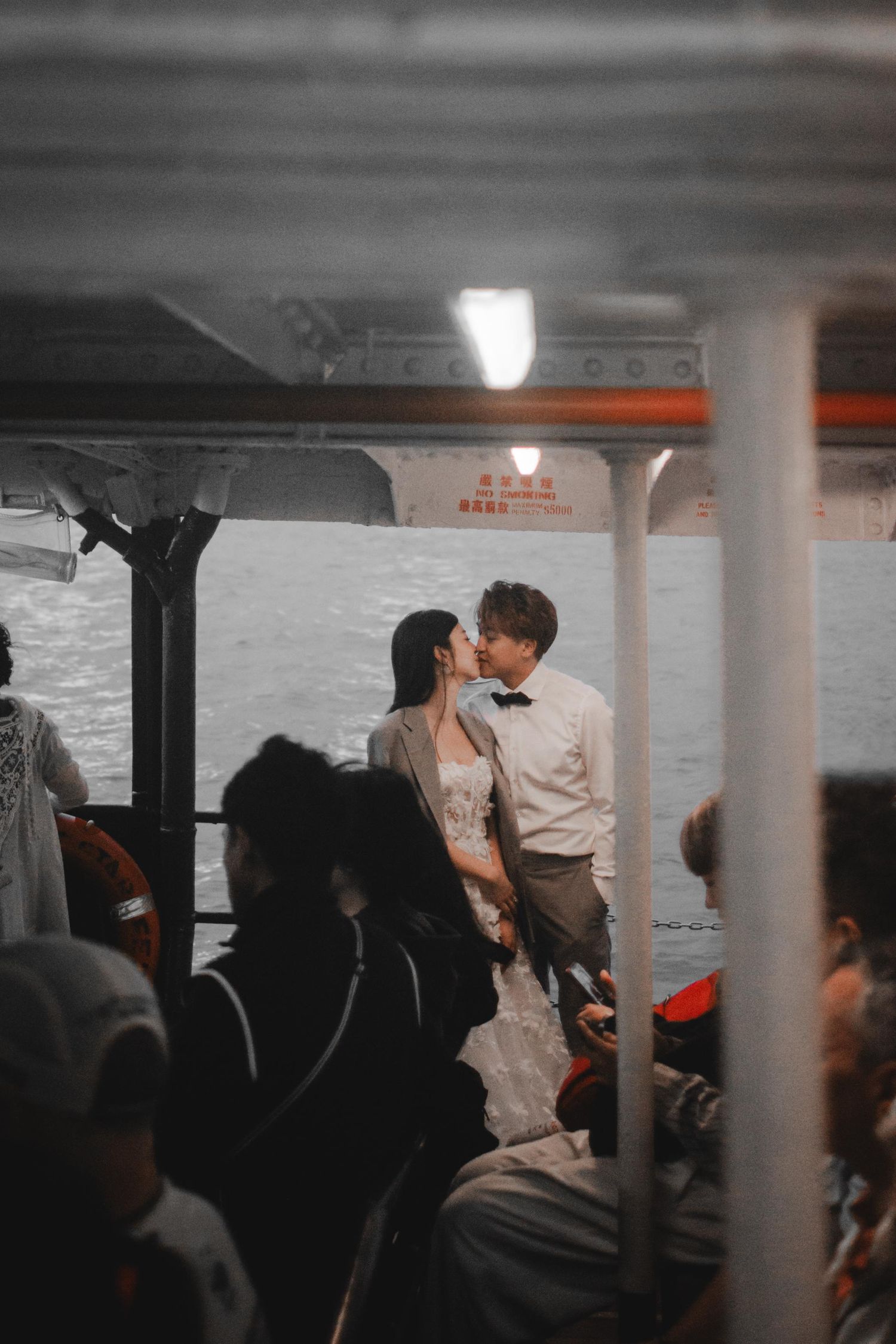 A wedding couple shares a romantic kiss on a ferry boat surrounded by passengers at night.