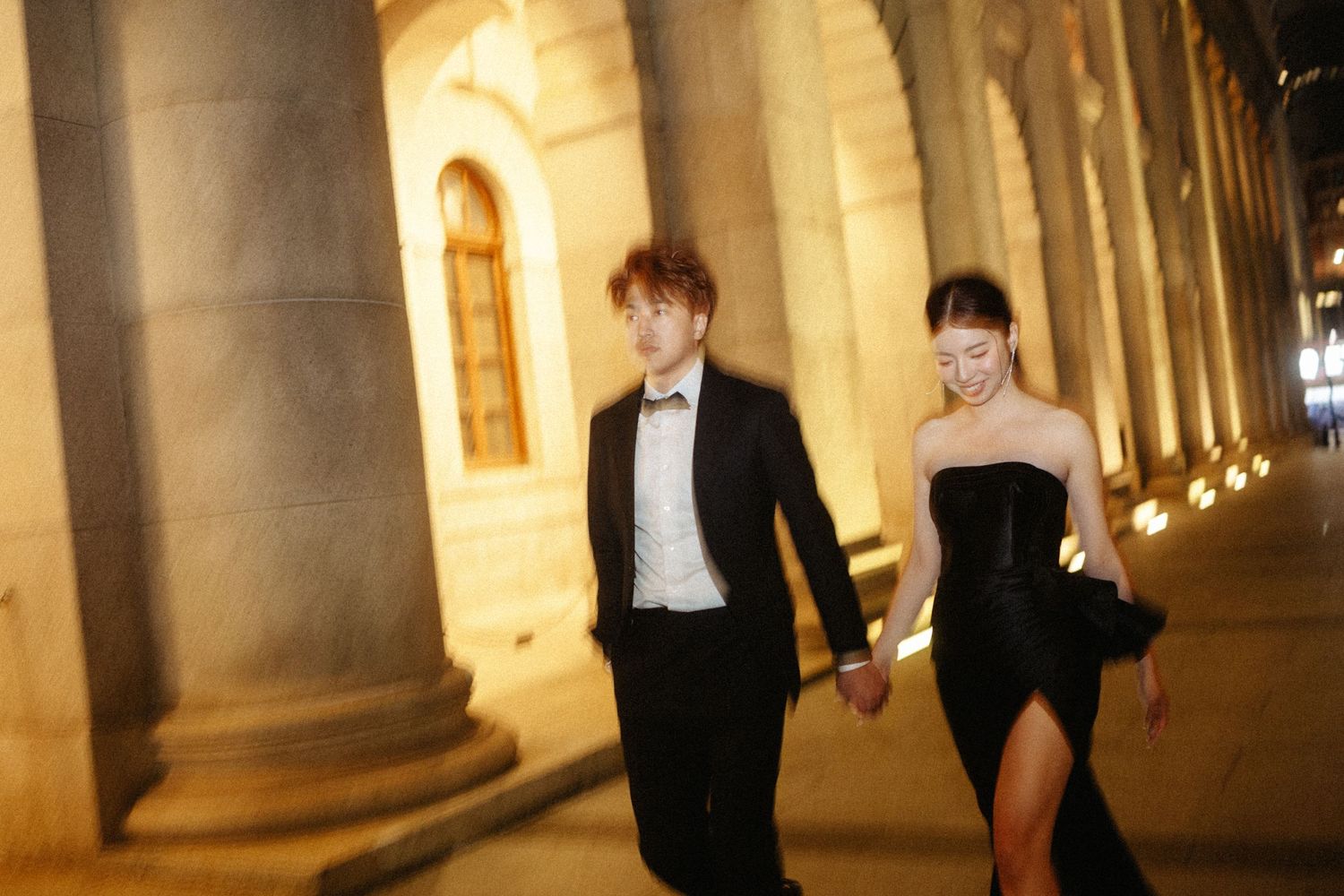 A couple in formal attire walks hand in hand past grand stone columns at night.