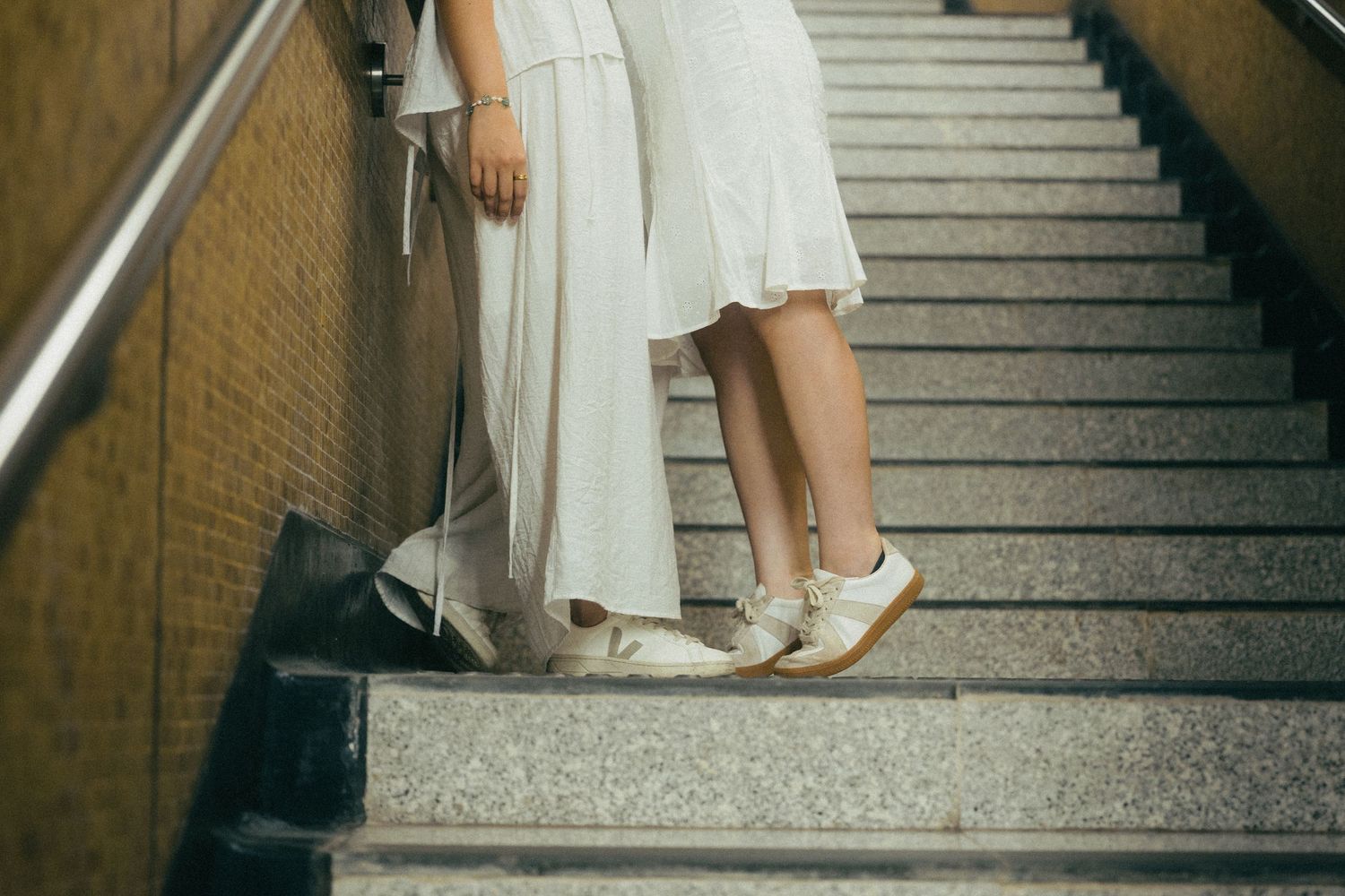 Two women in white dresses standing on stairs, one wearing white oxford shoes.