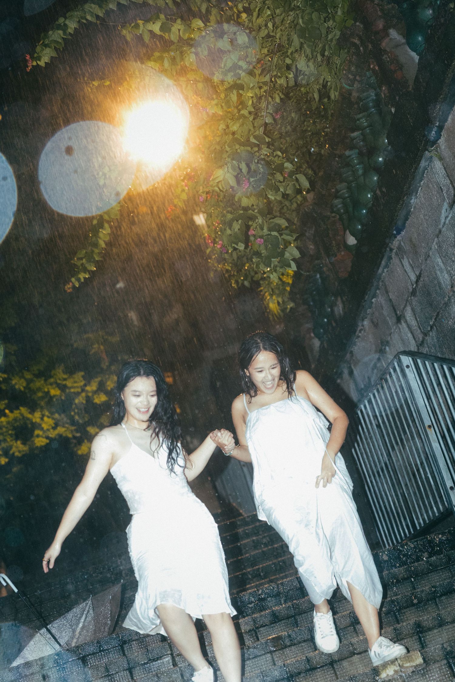 Two women in white dresses run hand-in-hand down steps in the rain under a glowing street lamp.