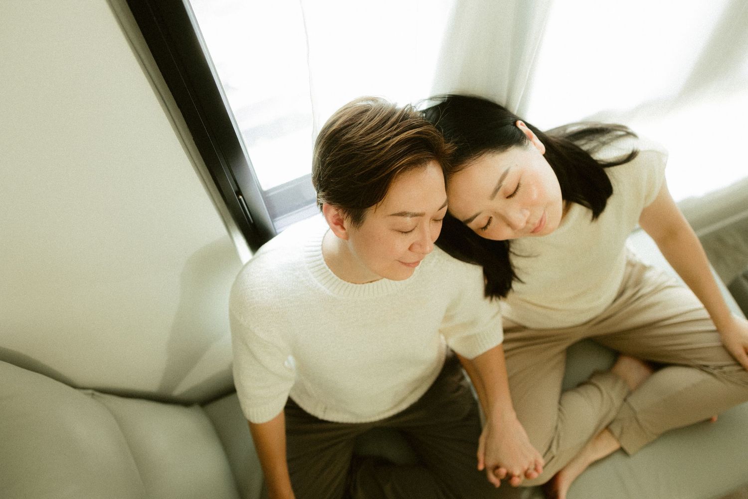 Asian couple holding hands while sitting closely together on a white sofa near window.