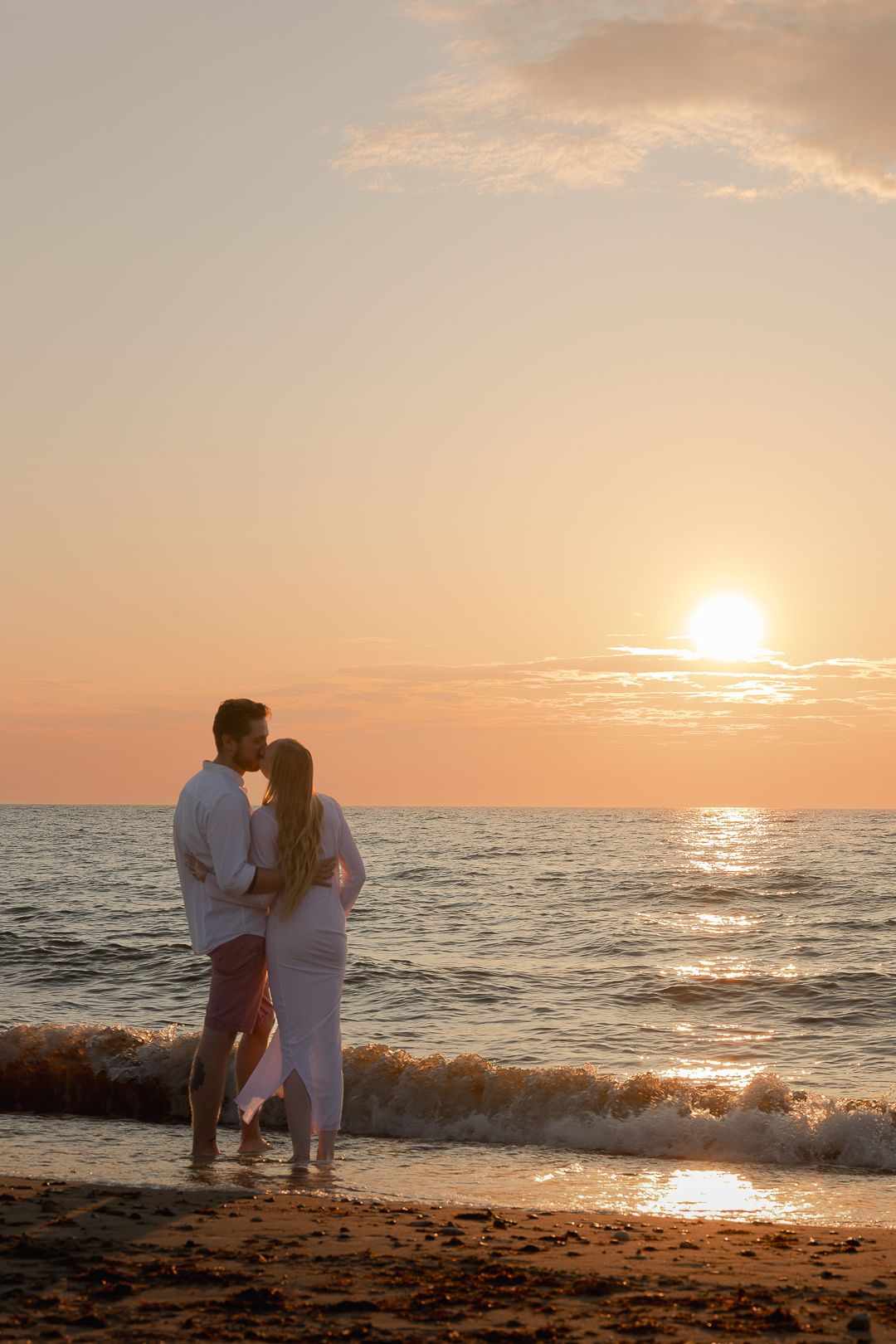A couple stands together at the water's edge on a sandy beach, embracing while watching a vibrant sunset.