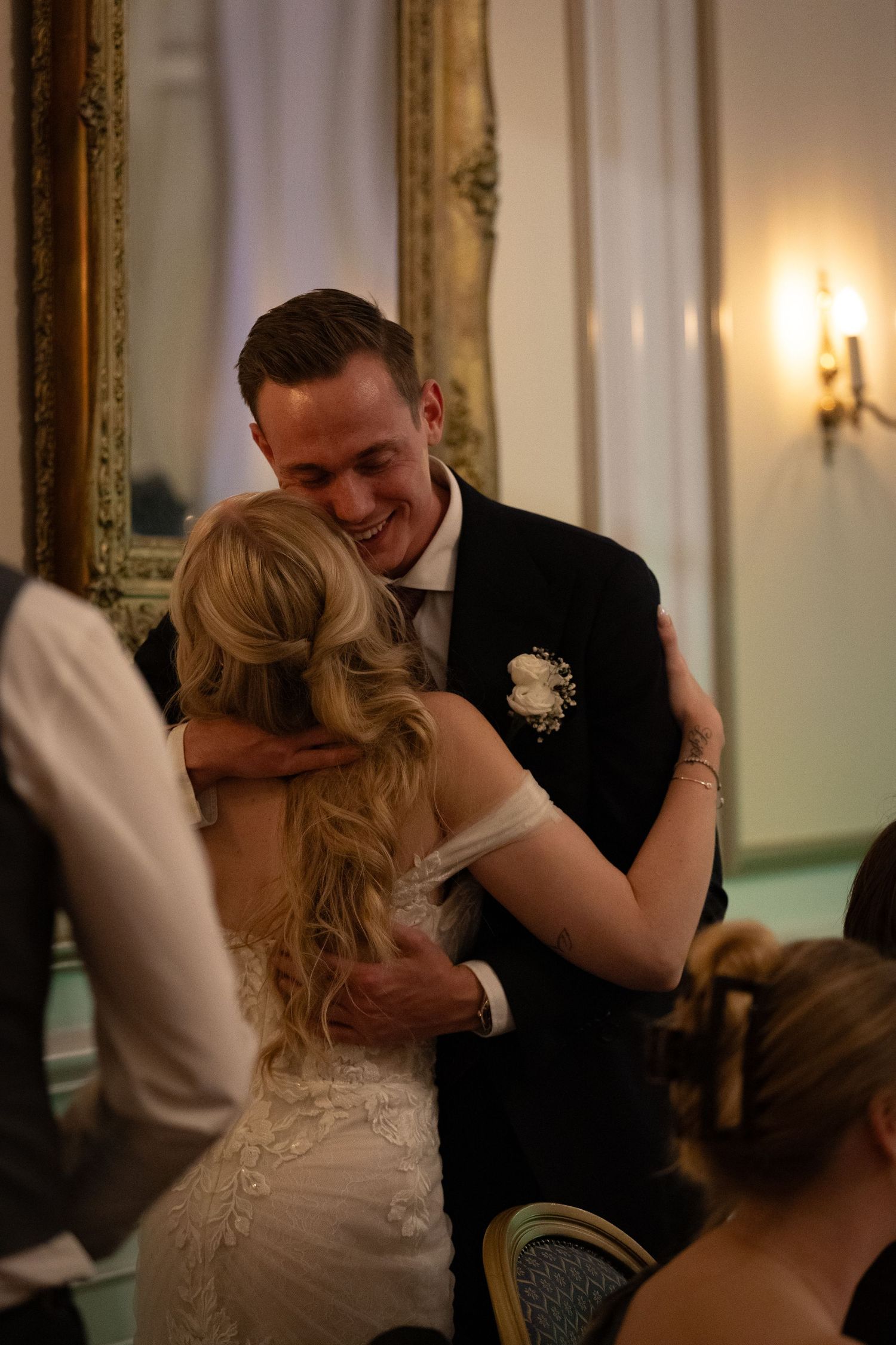 Smiling groom in a dark suit embracing his blonde bride in a lace wedding dress at their elegant reception.