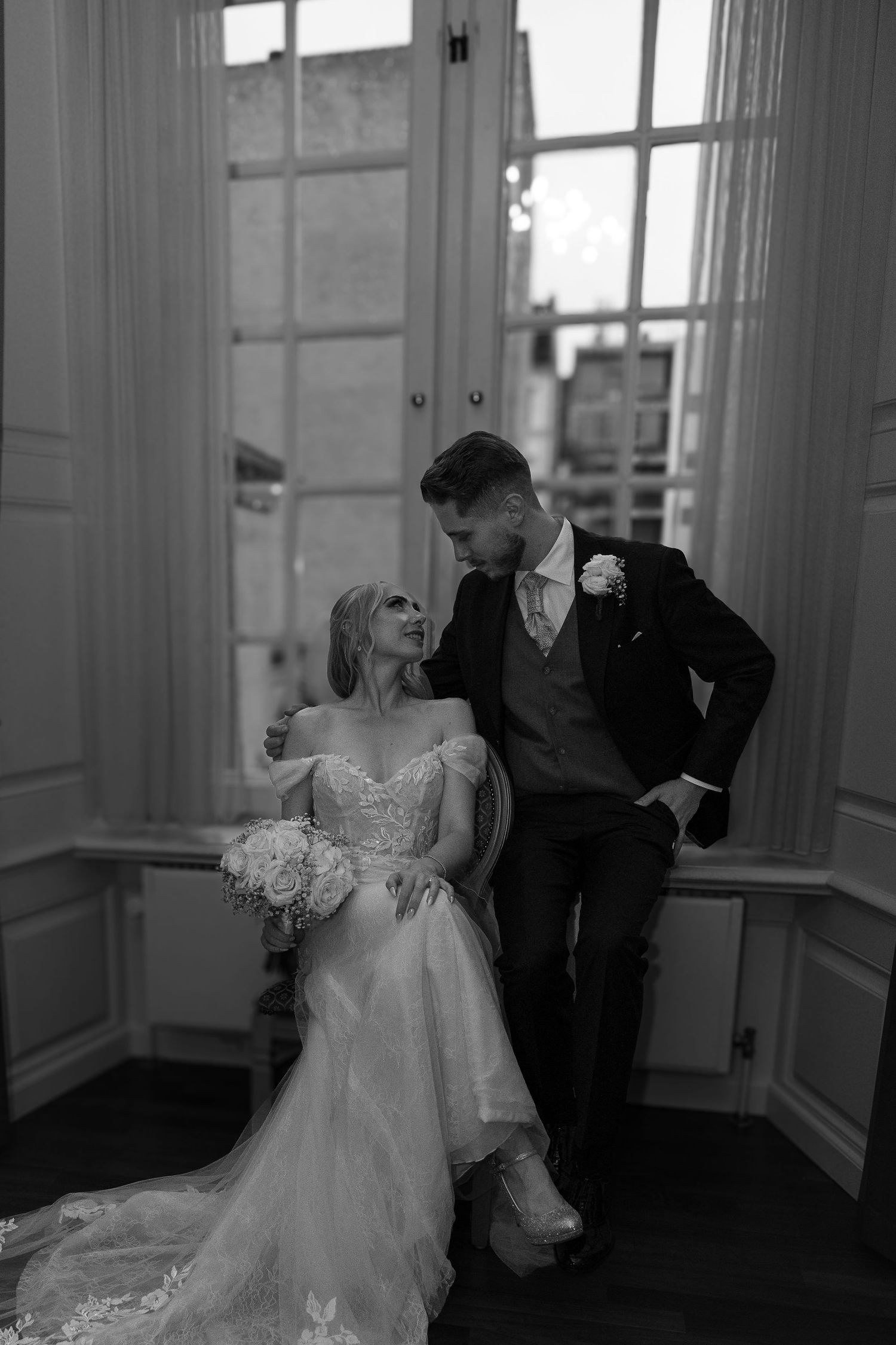 Bride and groom sharing loving glance by tall windows in elegant venue.