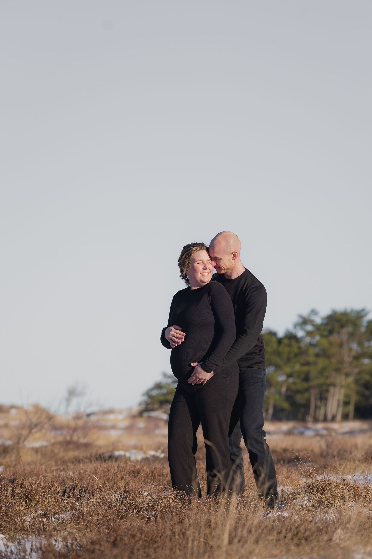 Couple in matching black outfits embracing outdoors in a snowy winter field, pregnant woman cradling her baby bump.