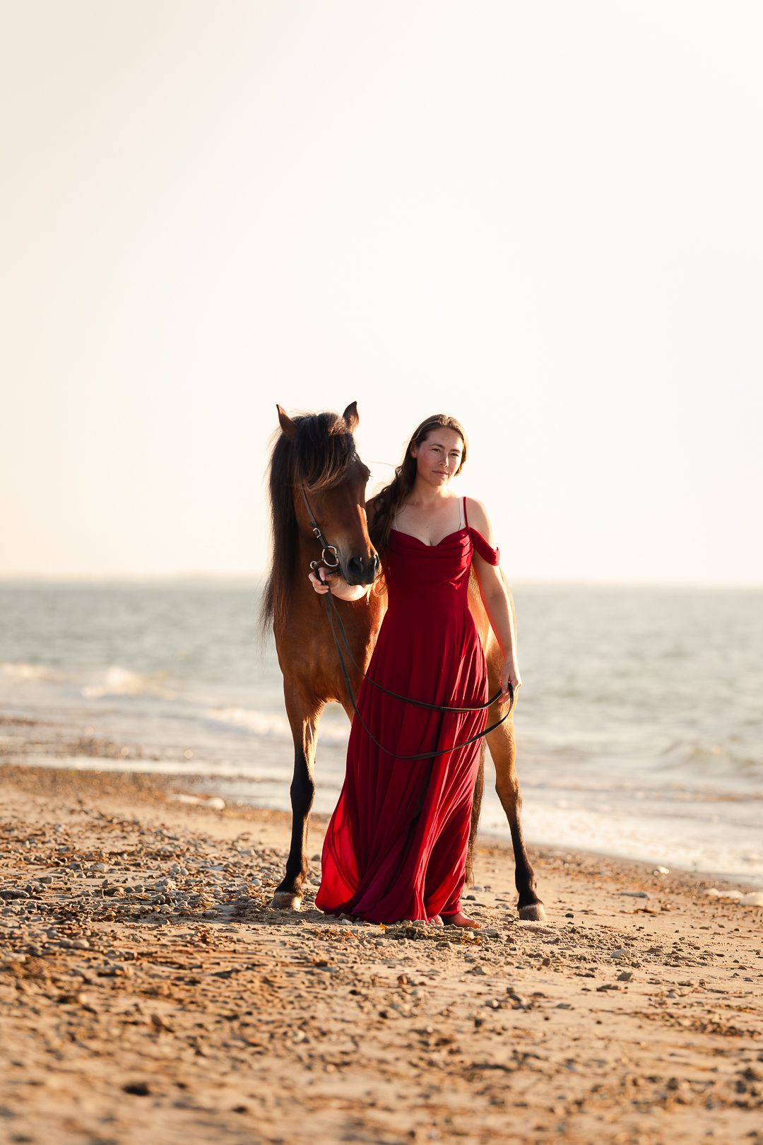 Smiling woman in red dress walking alongside brown horse on sunlit sandy beach.