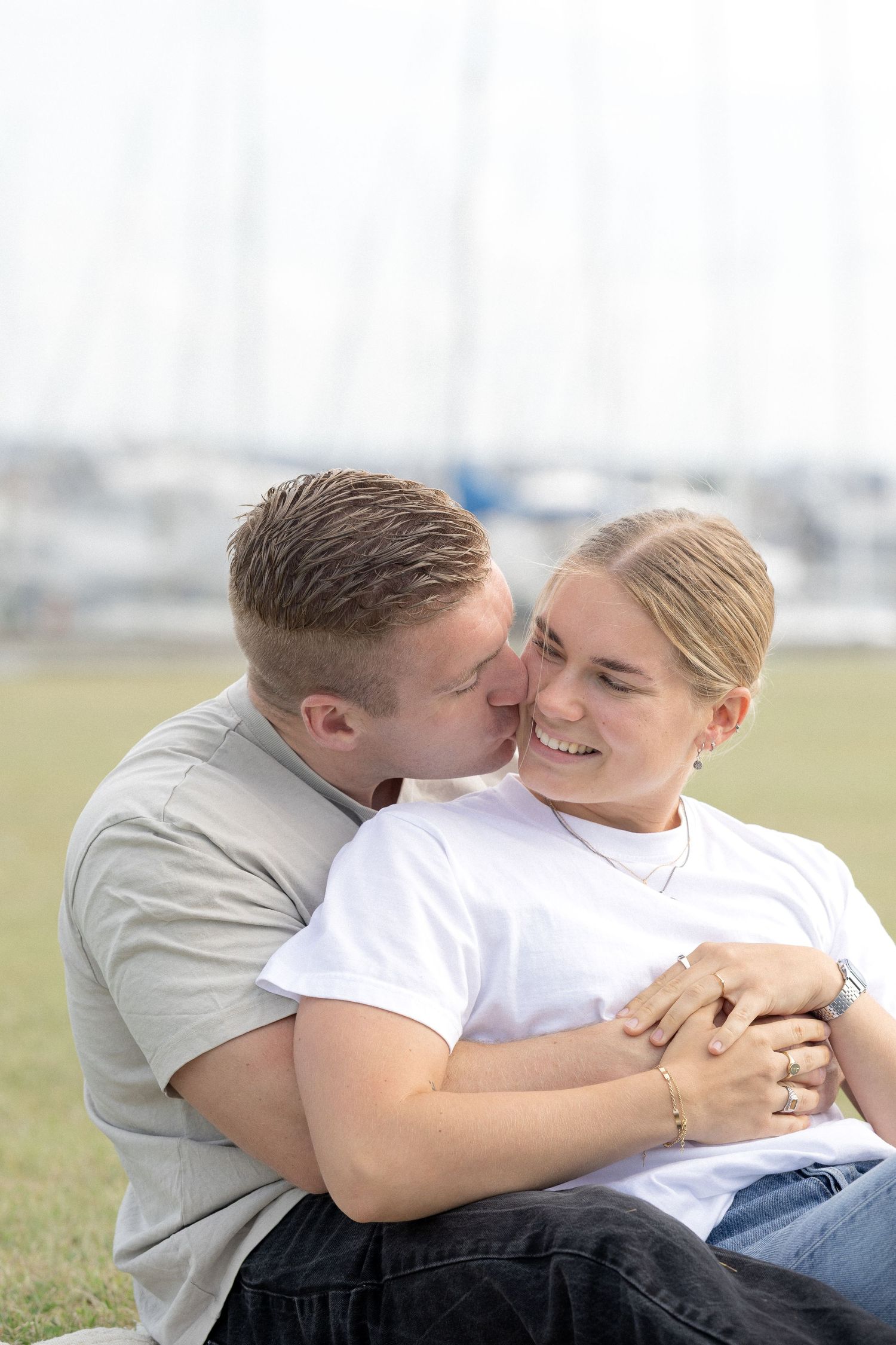 Man kissing smiling woman on cheek while embracing outdoors near a marina with sailboats.