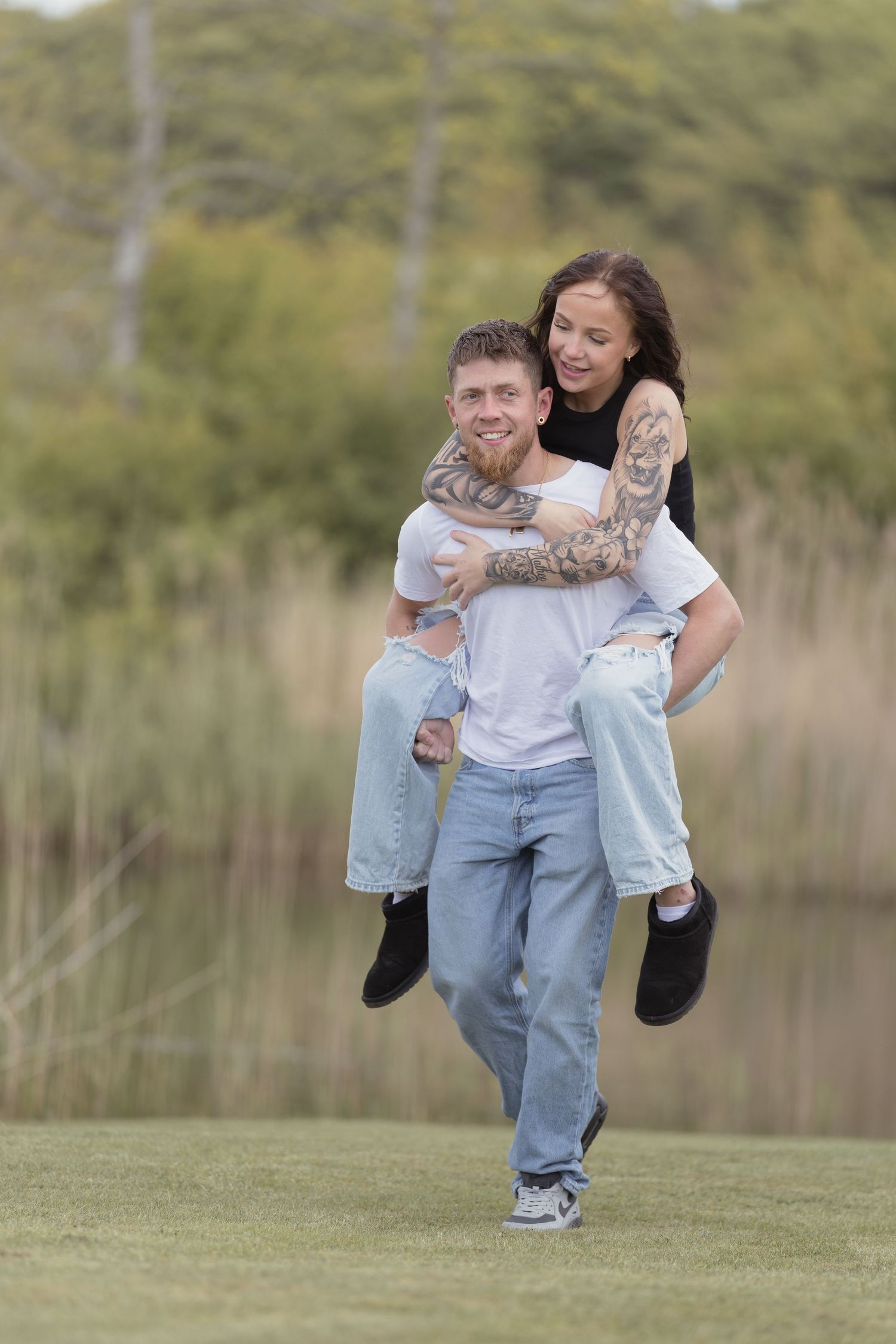 Tattooed couple laughing as man carries woman piggyback across a green lawn near a lake.