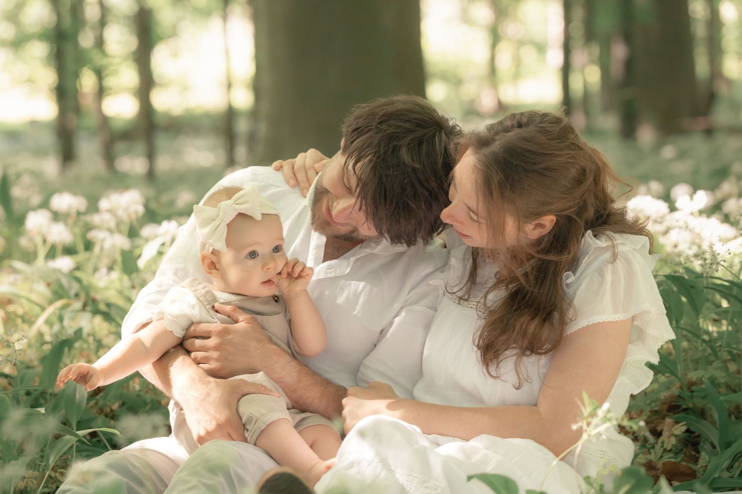 A happy family of three sits together in a sunlit forest surrounded by white flowers.