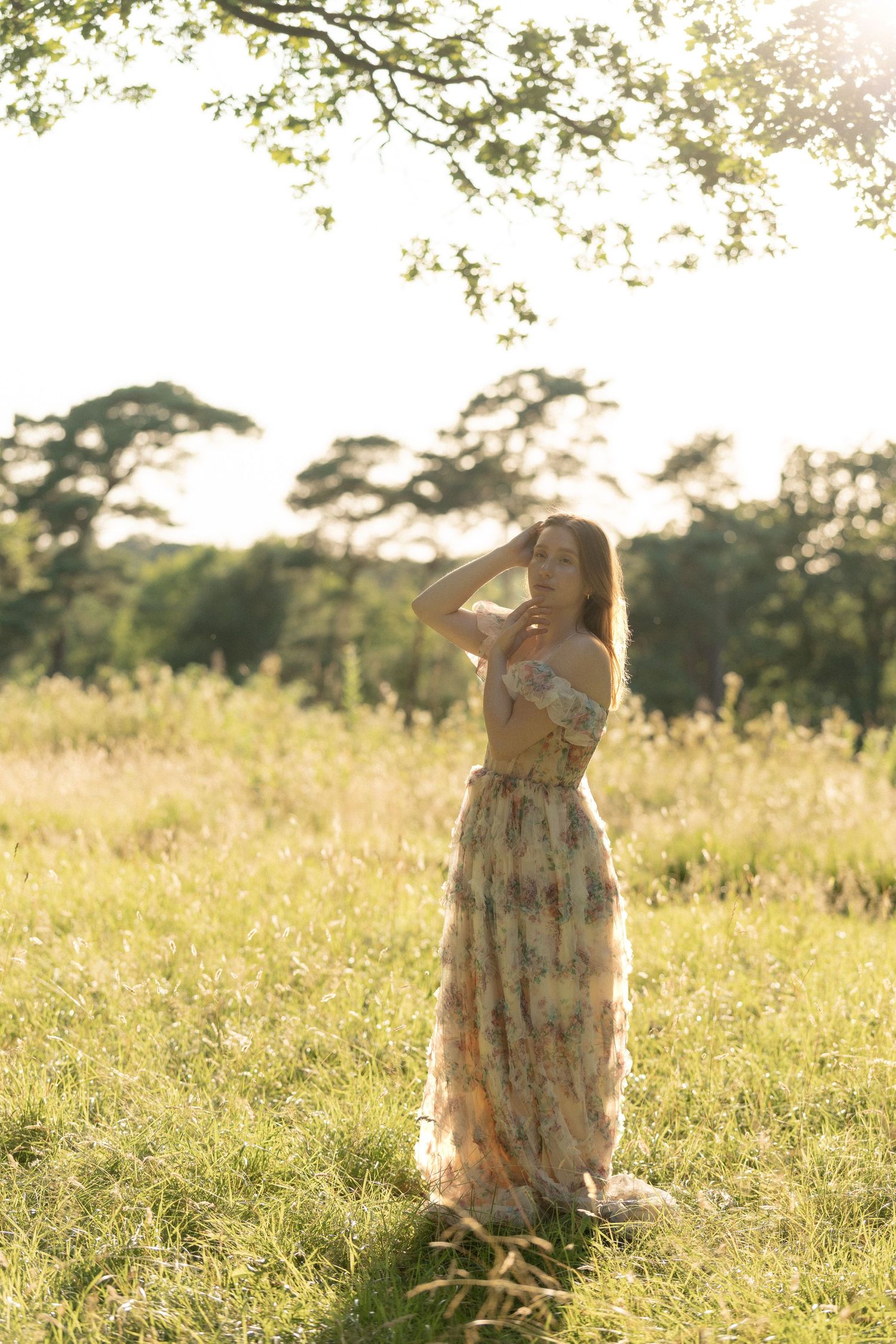 Woman in floral maxi dress standing in sunlit meadow with trees, golden hour light, hands raised near face.
