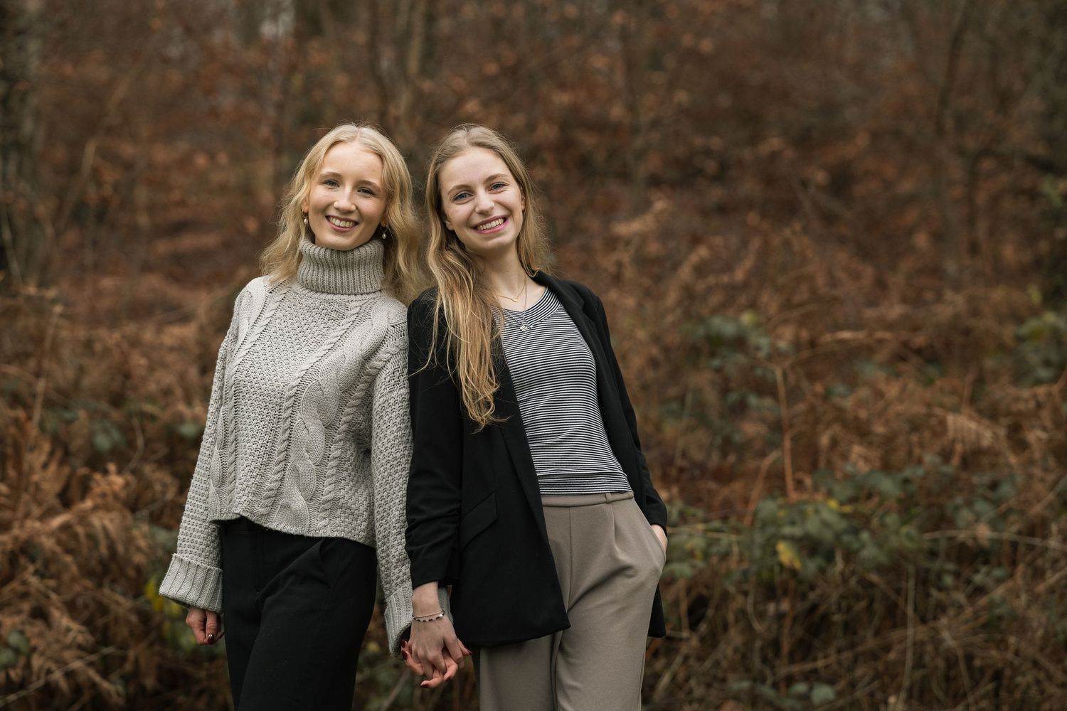 Two young blonde women holding hands and smiling in an autumn nature setting.