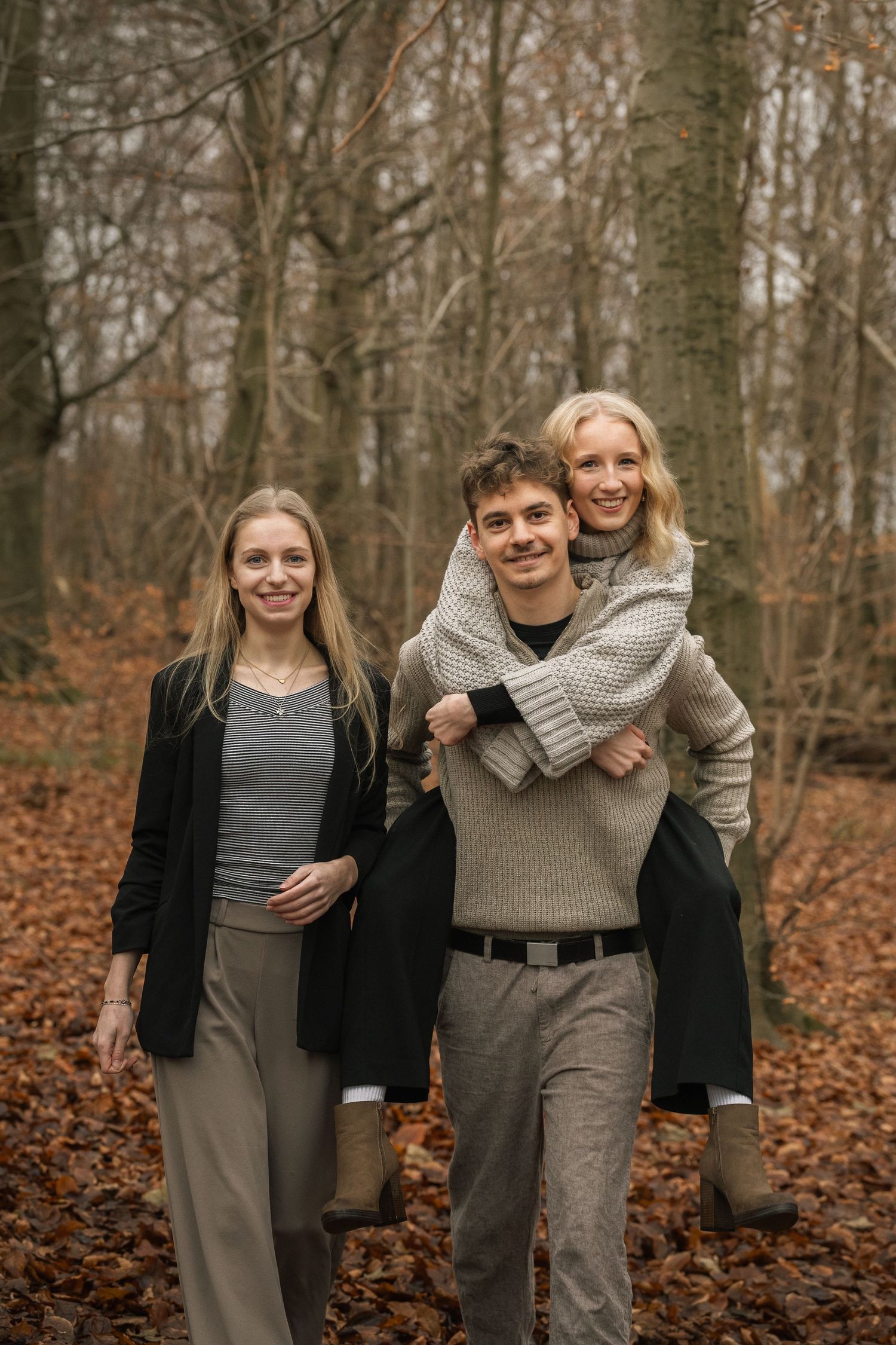 Three friends smile in an autumn forest as one woman gets a piggyback ride from a young man.
