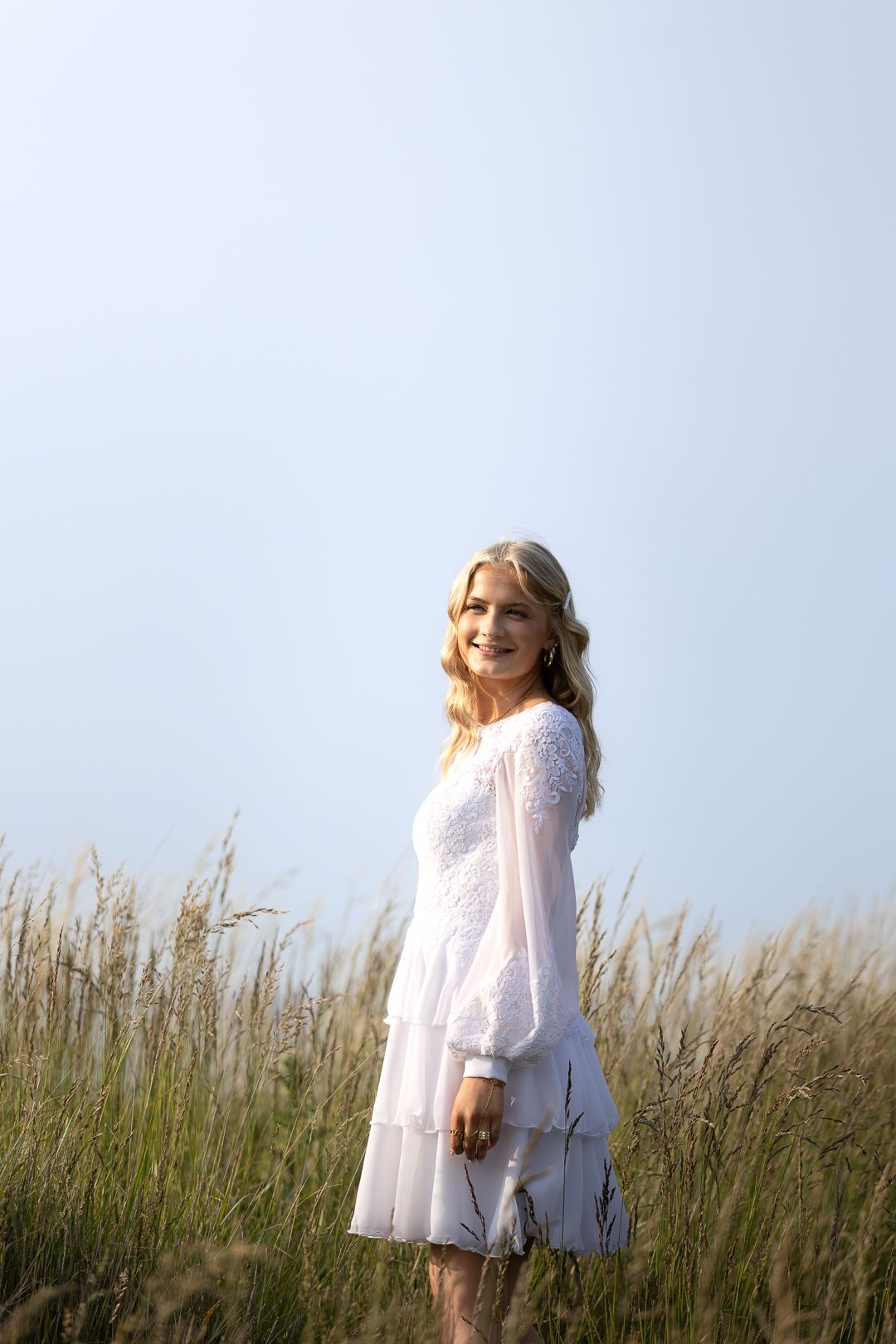 Smiling blonde woman in elegant white long-sleeve dress standing in sunlit tall grass meadow.