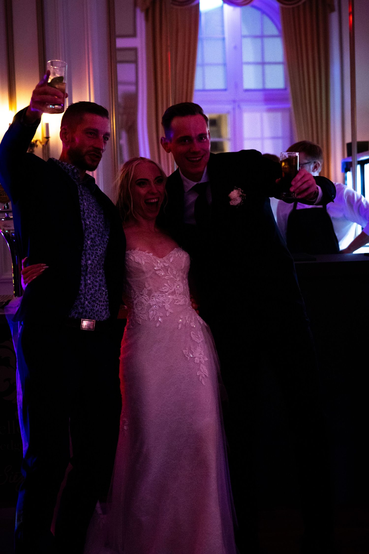 Bride in white gown celebrates with two suited guests raising drinks under warm purple wedding reception lighting.
