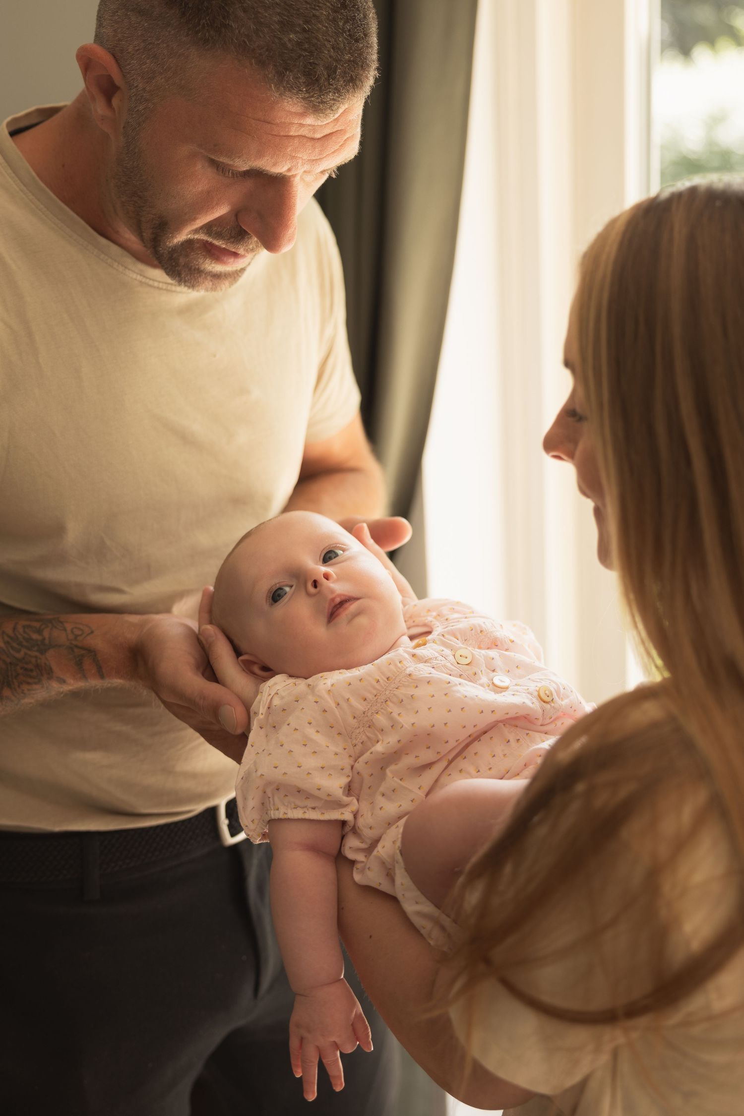 A tattooed father and mother lovingly holding their newborn baby in a pink onesie near a sunlit window.