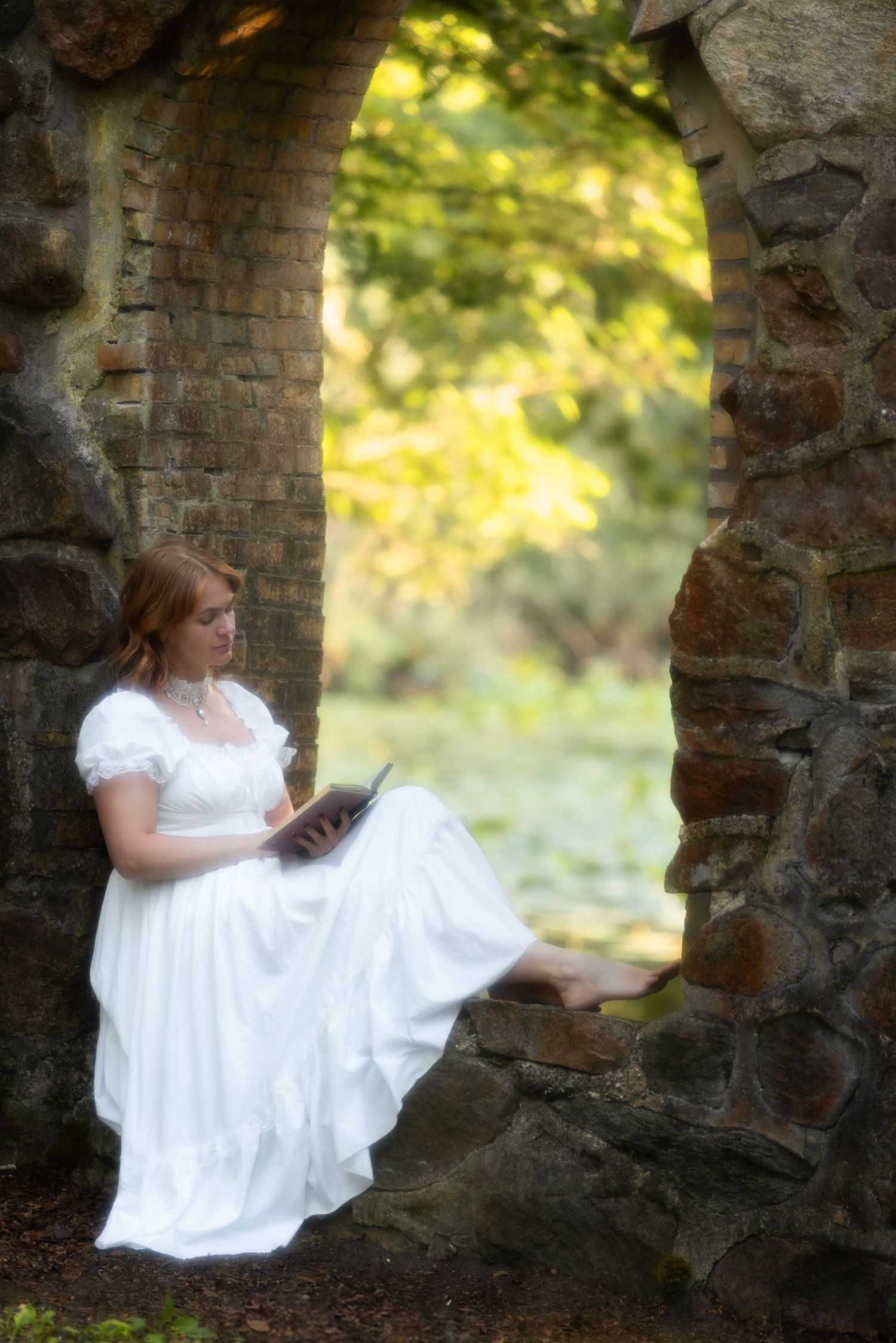 Young woman in white vintage dress reading book in stone archway surrounded by greenery.