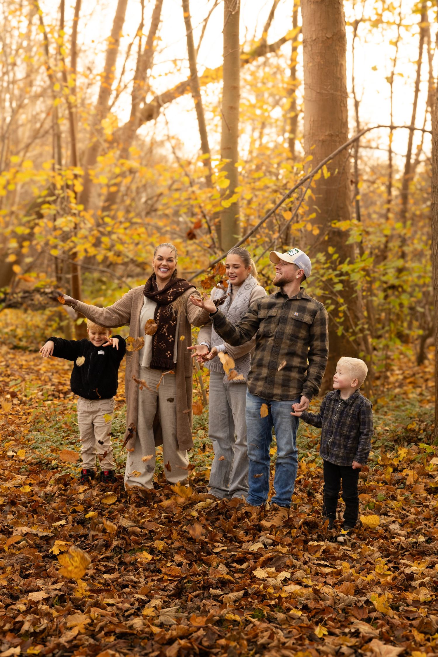 Joyful family of five tossing autumn leaves in a sunlit forest with golden fall foliage.