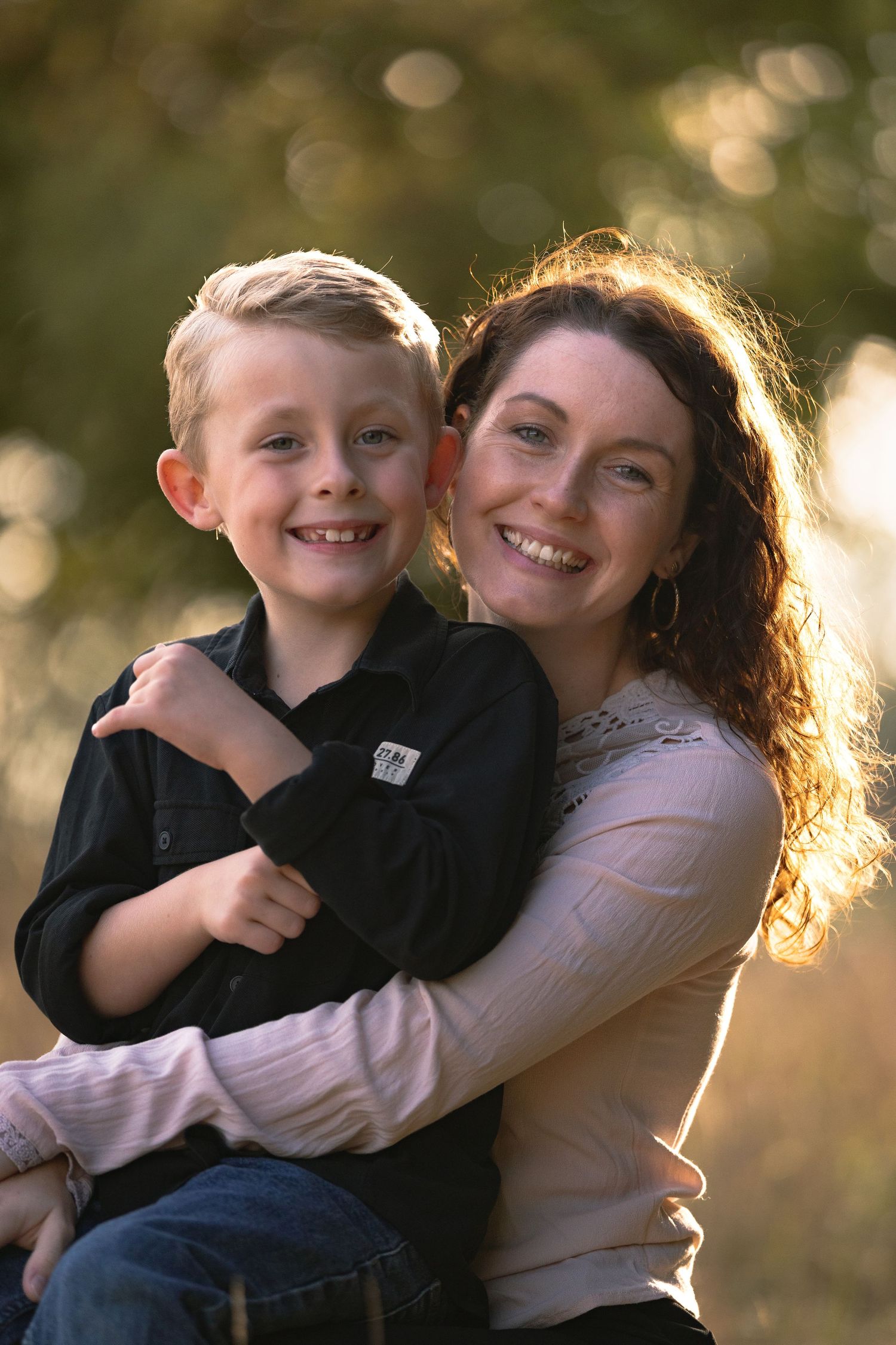 Smiling mother hugging her young son outdoors in warm golden sunlight.