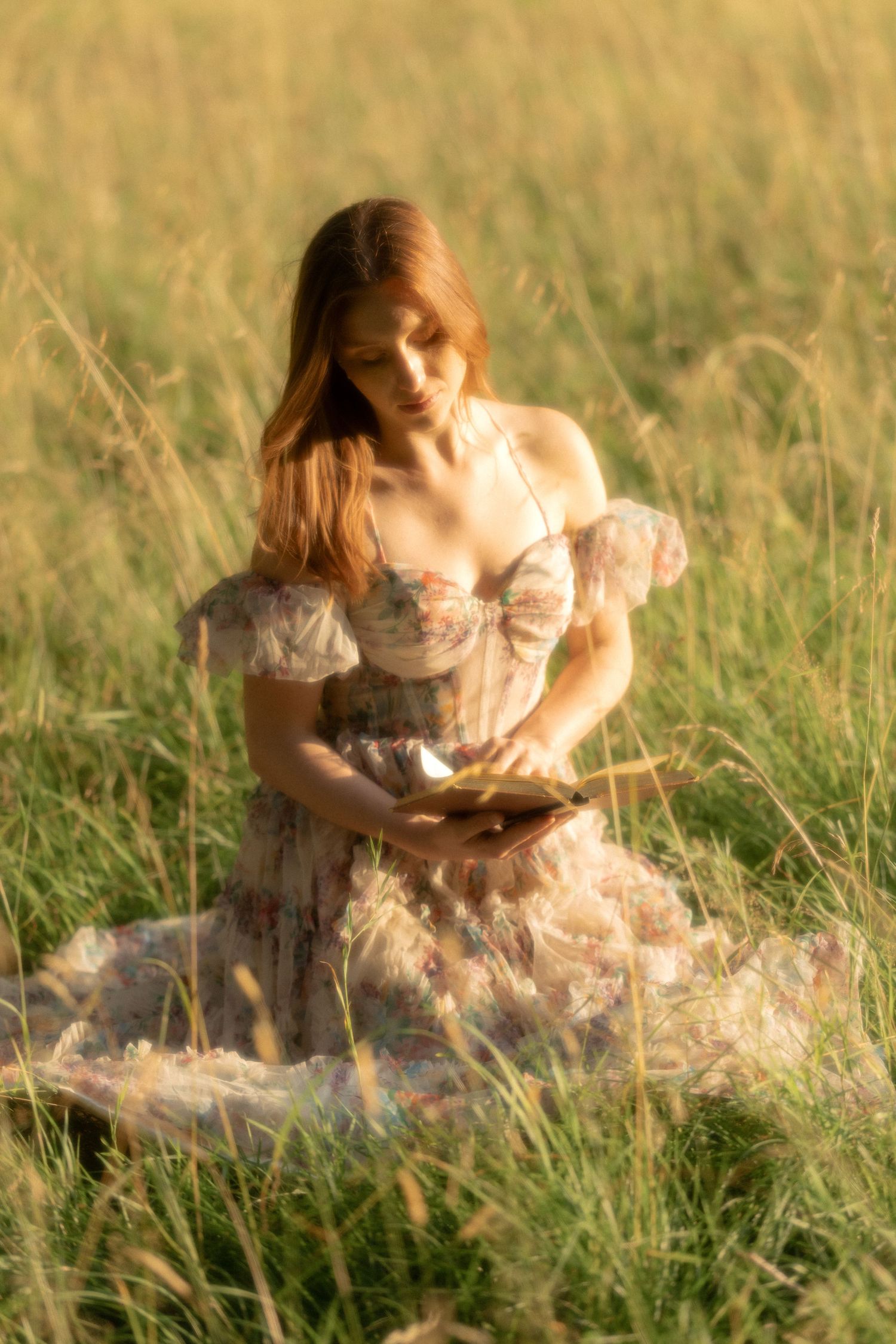 Young woman in floral corset dress reading book while sitting in golden sunlit grass field.