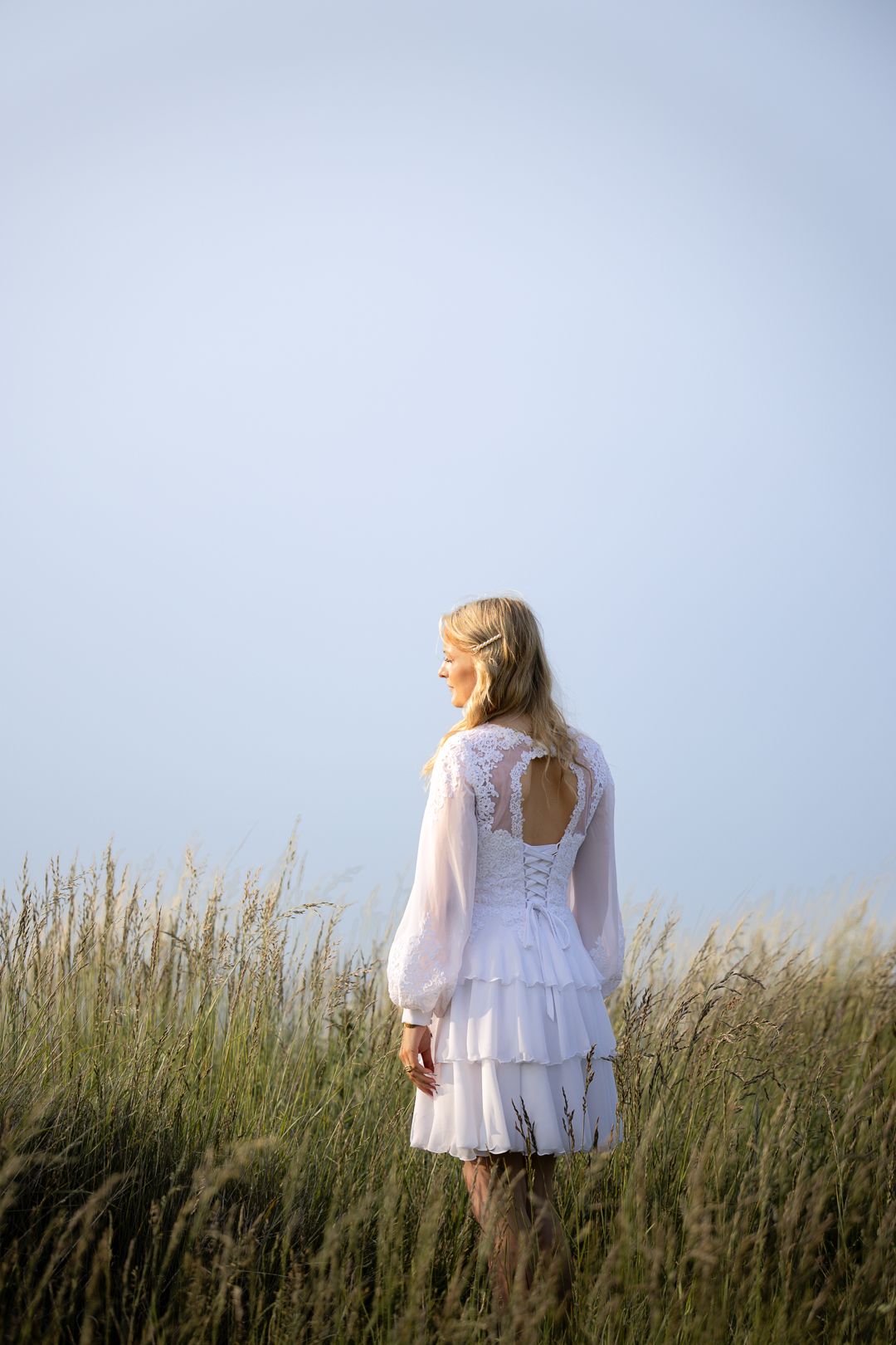 Blonde girl in white lace dress with open back standing in golden grass field.