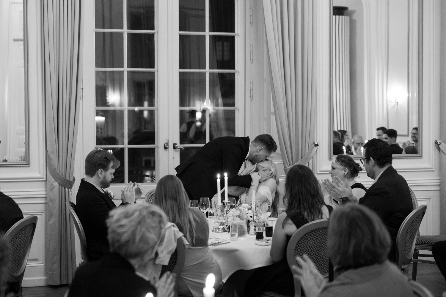 Black and white wedding reception dinner scene with guests seated at candlelit round tables in elegant venue.