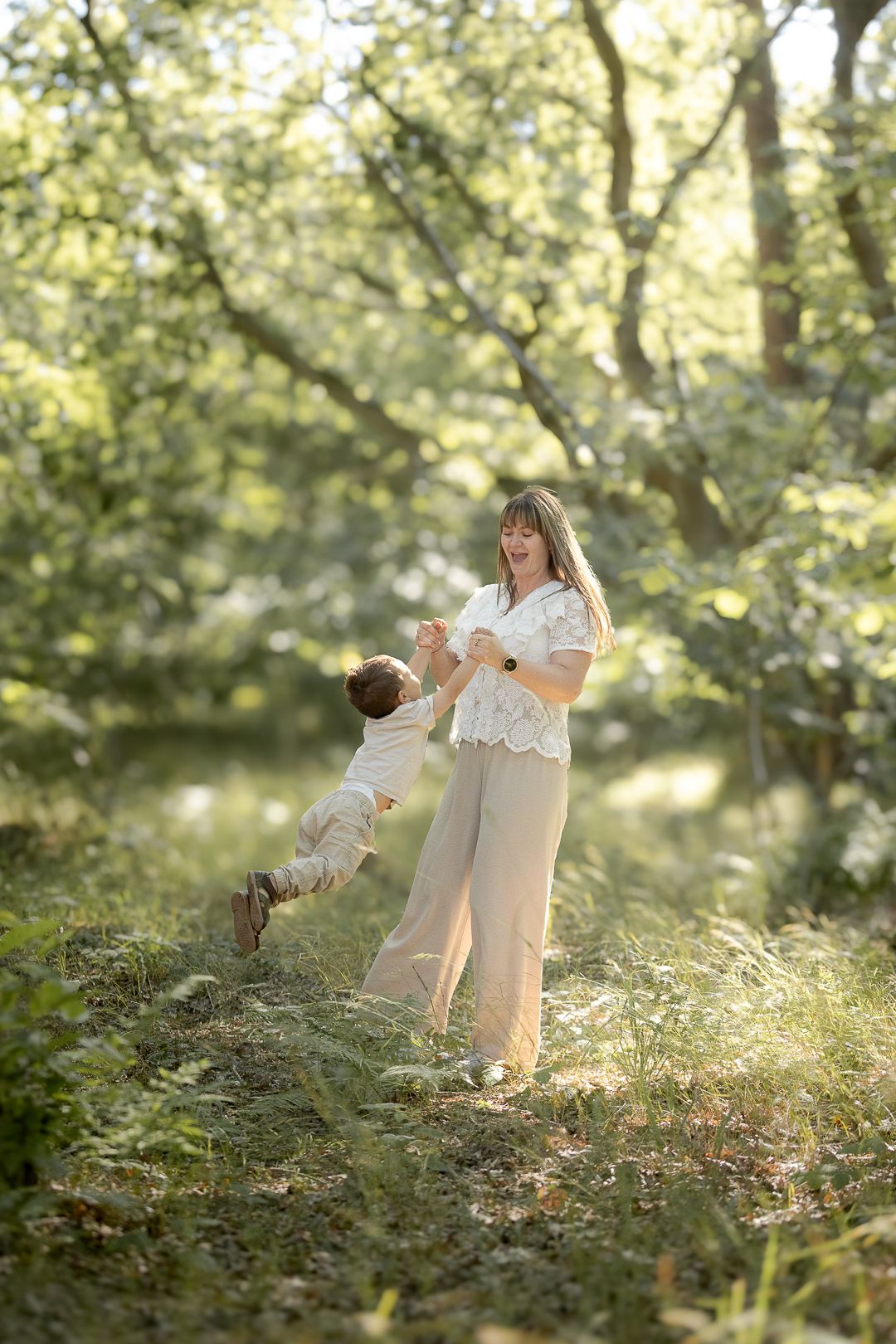 Mother joyfully spinning her toddler son in a sunlit green forest clearing.