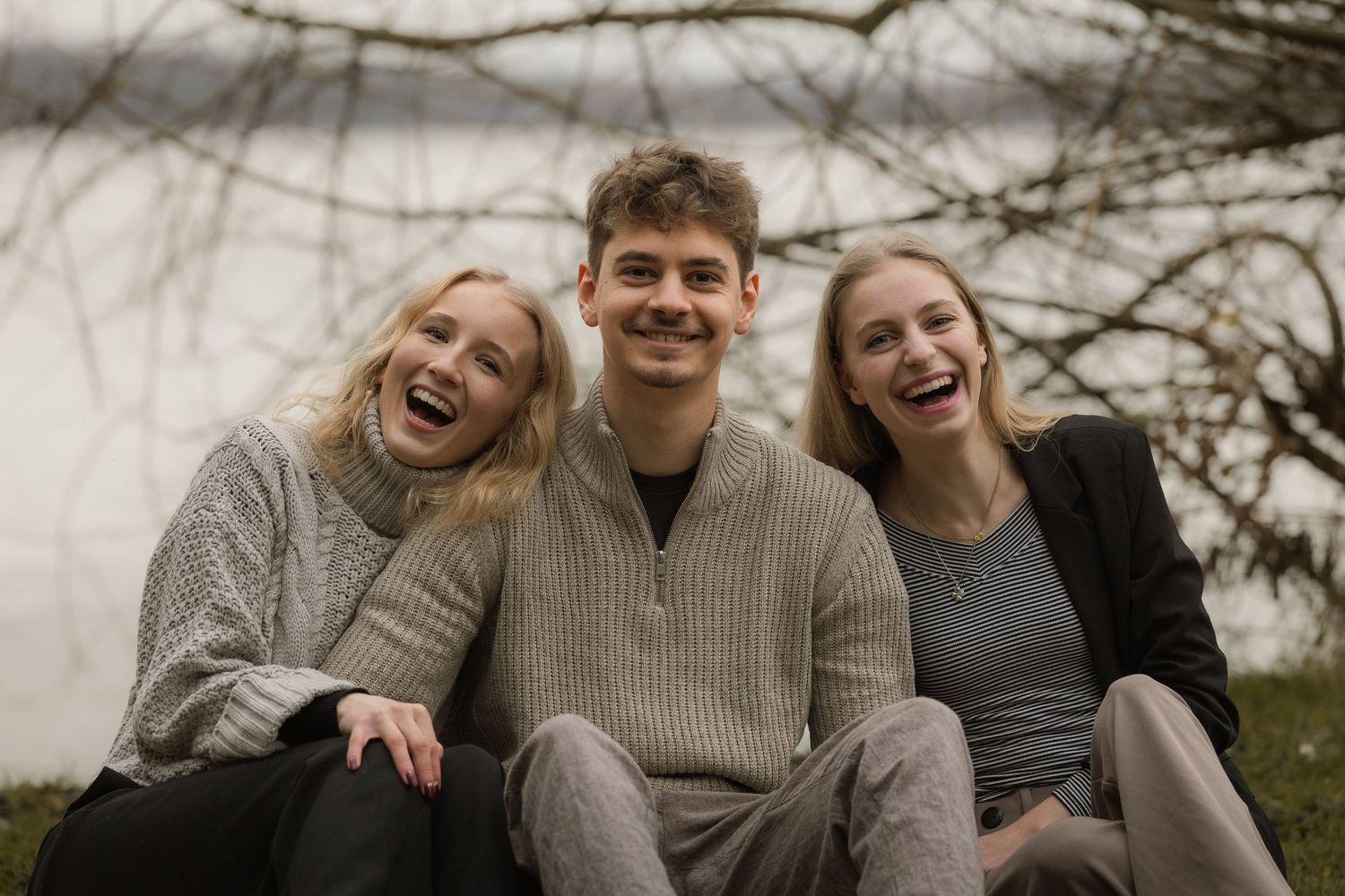 Three laughing young adults sit closely together outdoors near a lake on a cloudy day.